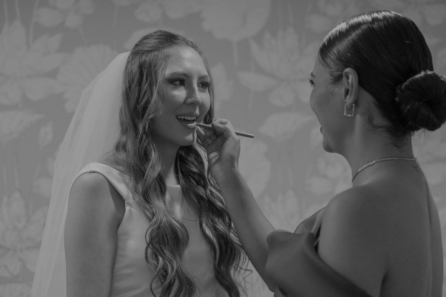Bridesmaid applying makeup to a smiling bride in a wedding veil before the ceremony.
