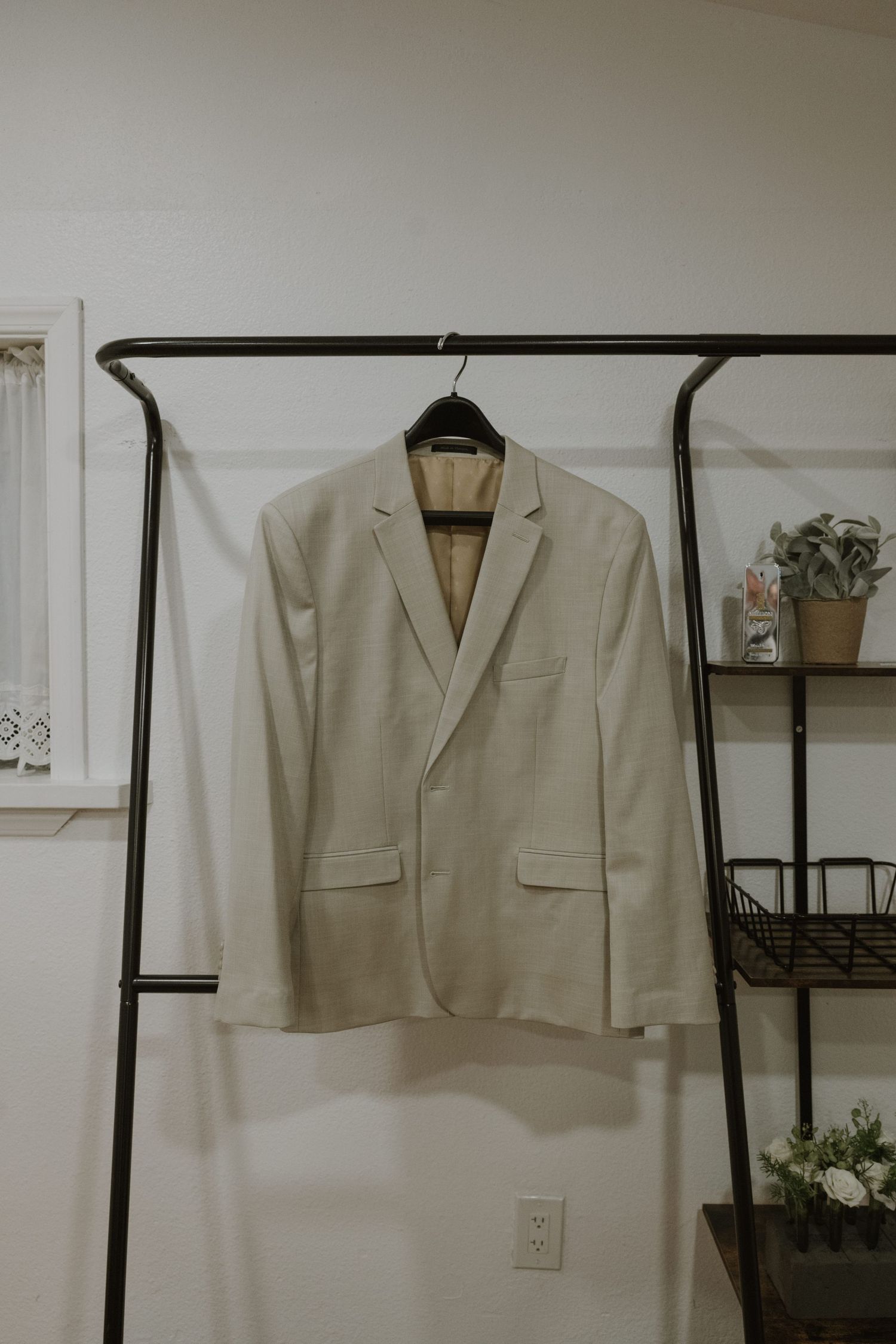 Beige blazer jacket hanging on a black hanger on a clothing rack against a white wall.