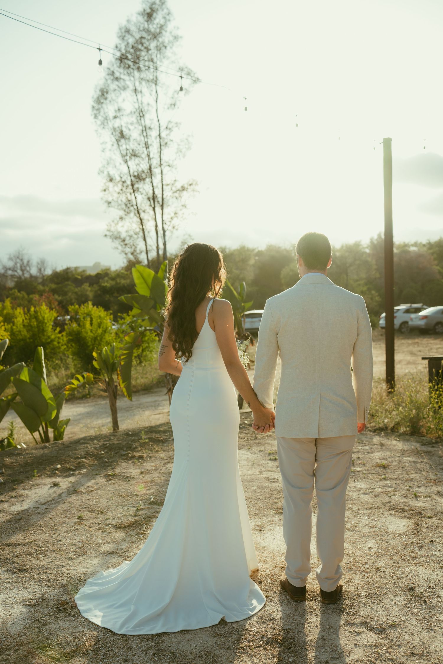 Bride and groom holding hands from behind at sunset, wearing white gown and beige suit outdoors.