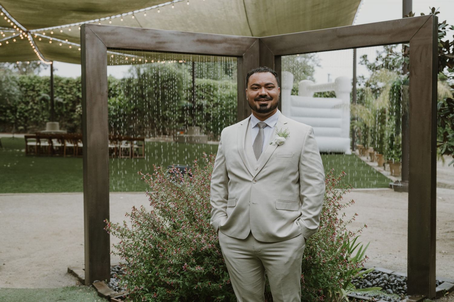 Groom in light gray suit smiling under wooden pergola at outdoor wedding venue with lush greenery backdrop.