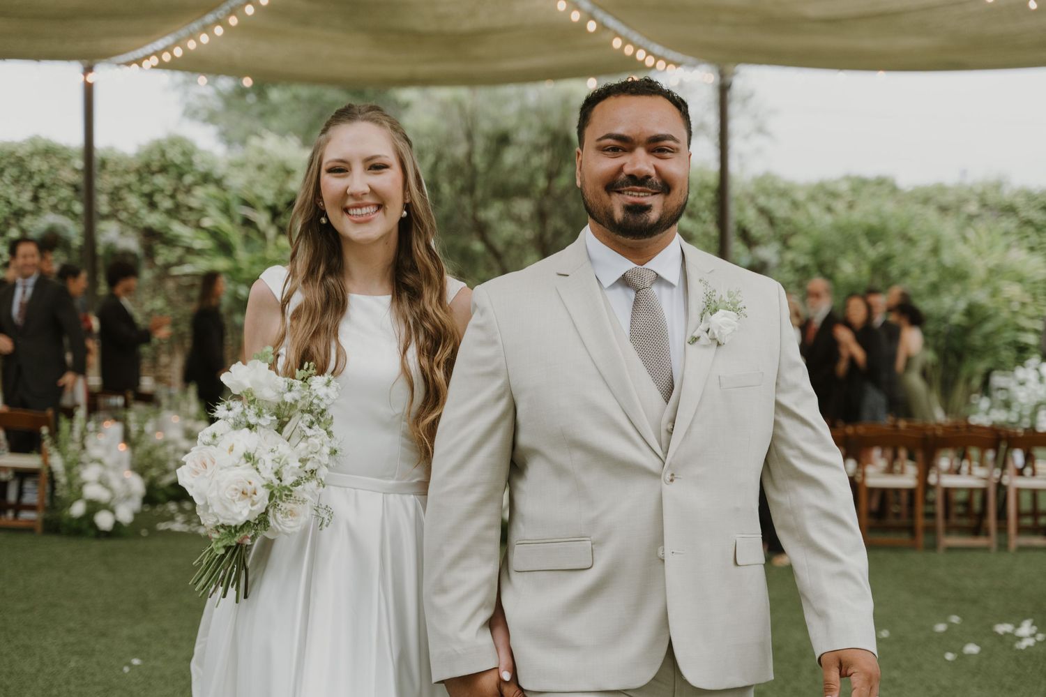 Newlywed couple smiling after outdoor wedding ceremony, bride holding white bouquet in elegant venue.