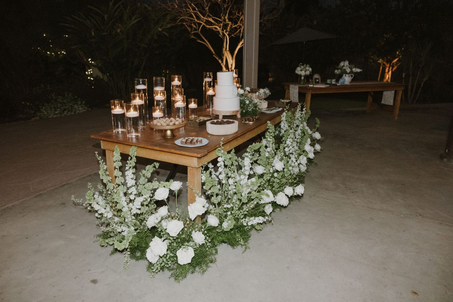 Rustic wooden sweetheart table adorned with white flowers, greenery, and glowing candles at an outdoor evening wedding.
