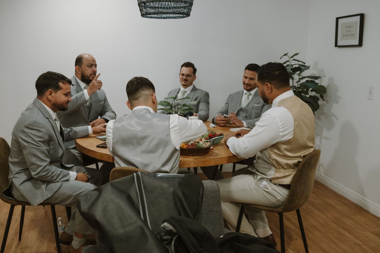 Groomsmen in gray suits gathered around a round wooden table laughing and talking before a wedding ceremony.