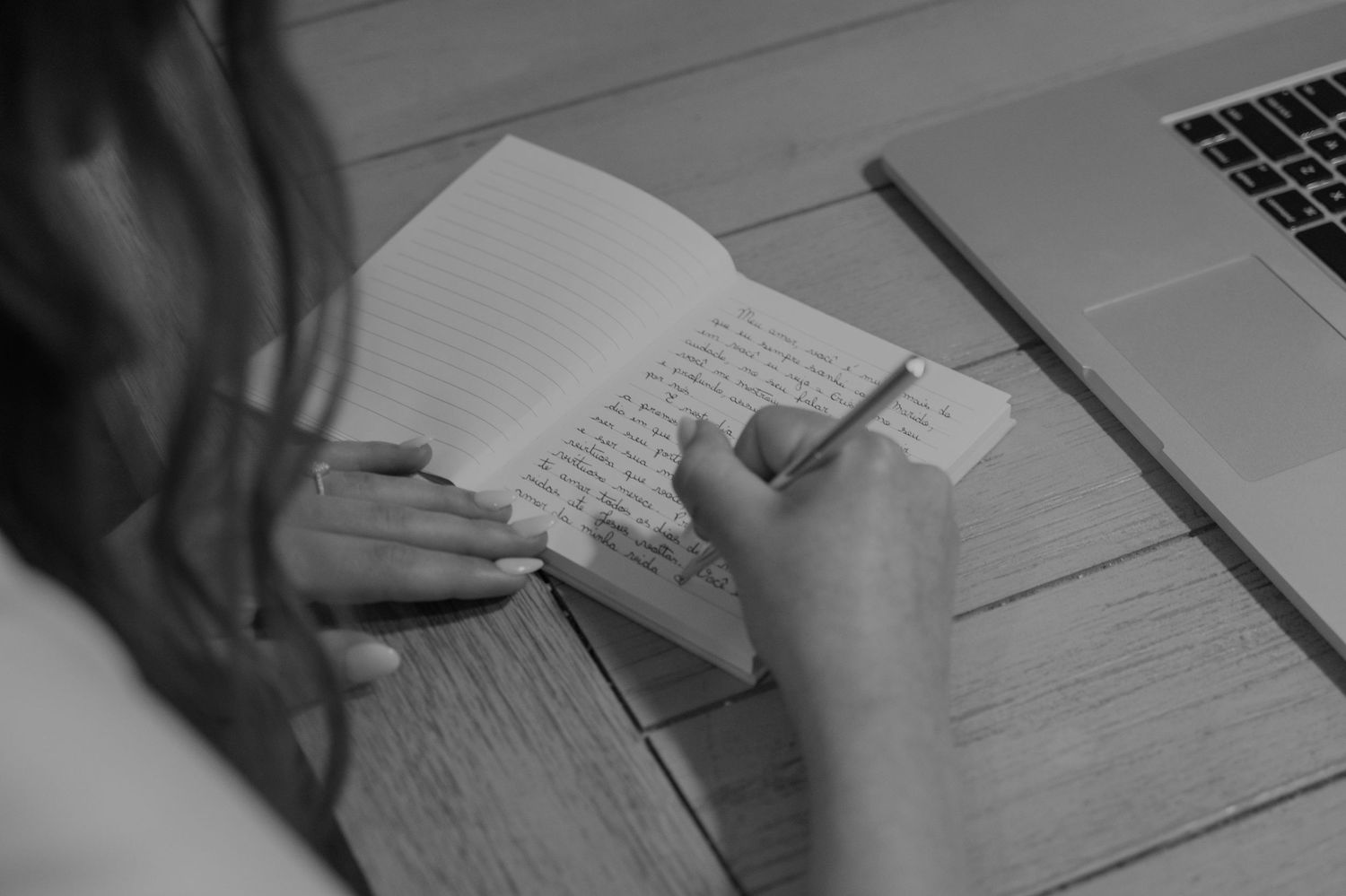 Woman writing in a notebook at a wooden desk with a laptop in the background, black and white photo.