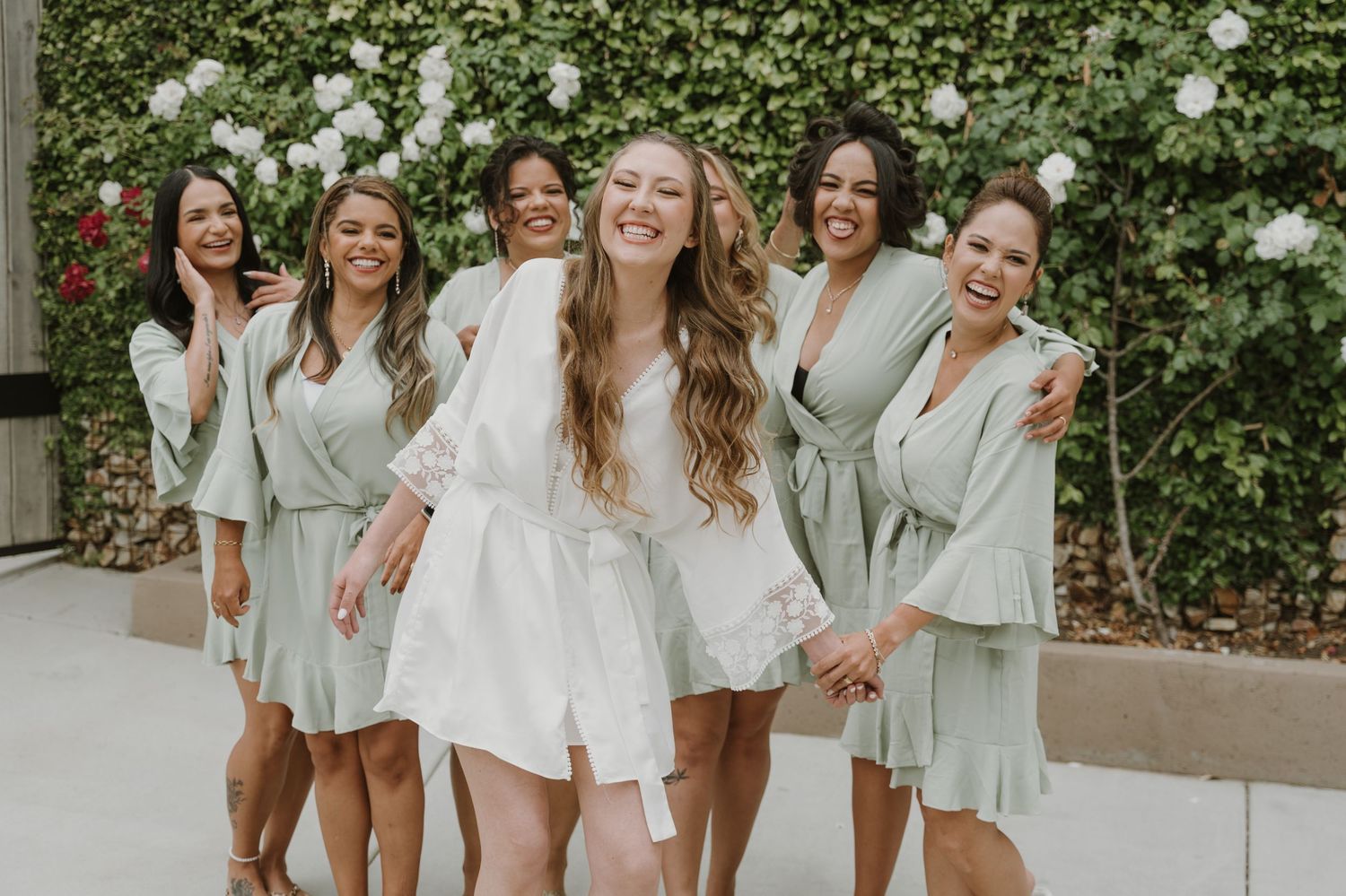 Smiling bride in white lace robe surrounded by laughing bridesmaids in sage green robes near floral wall.