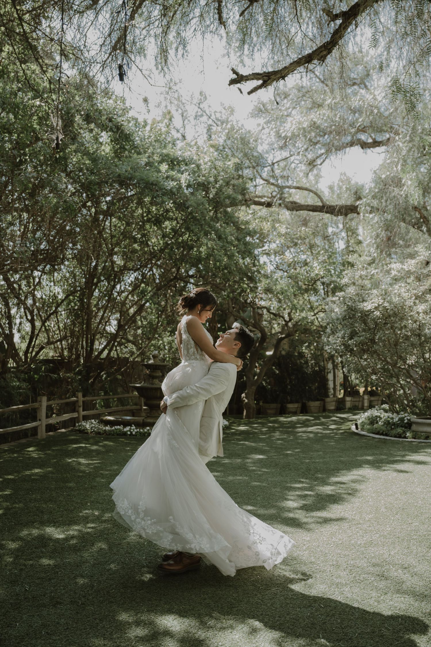 Groom lifts bride in white wedding gown outdoors among lush green trees on a sunny day.