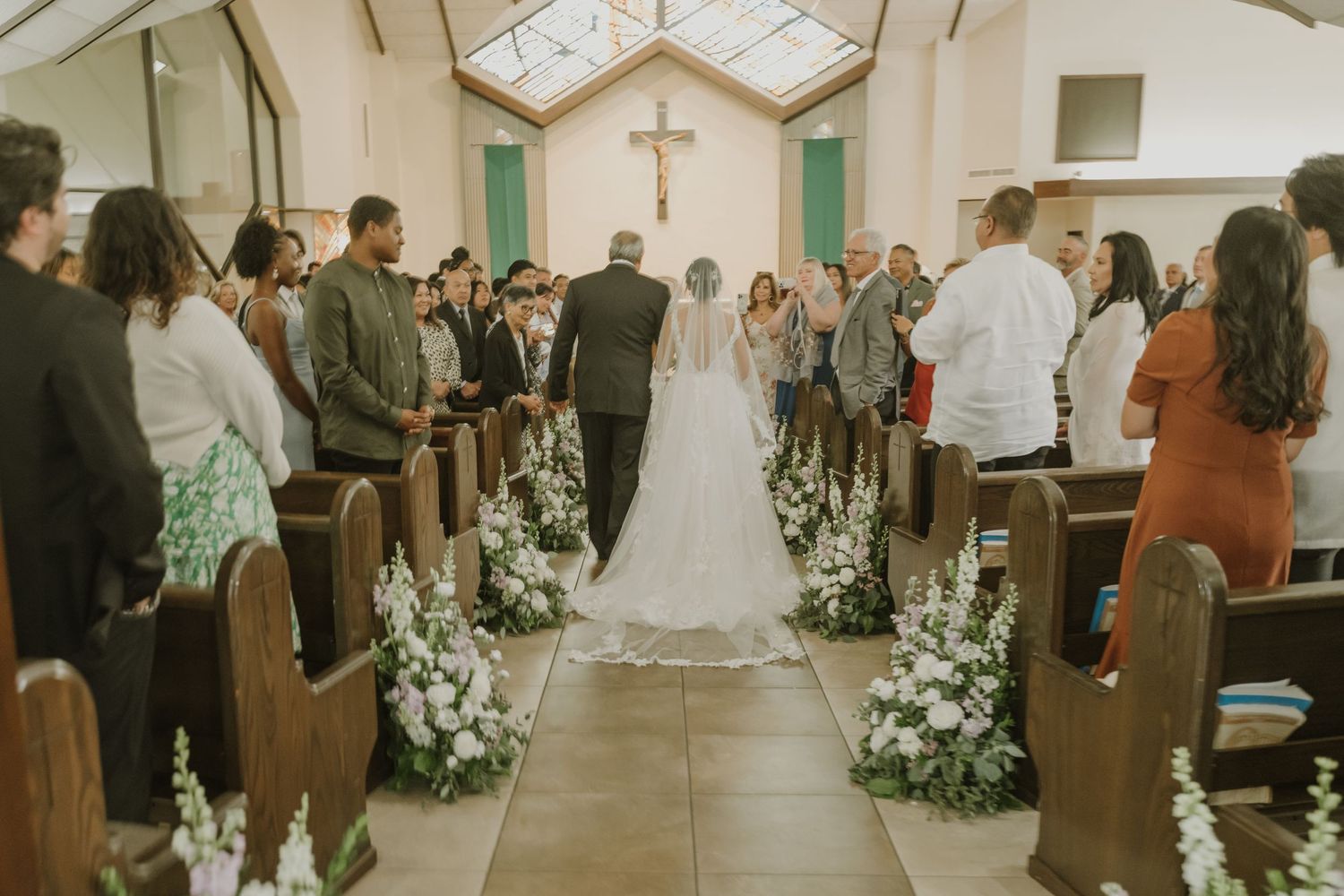 Bride walks down flower-lined church aisle with escort toward altar cross during wedding ceremony.