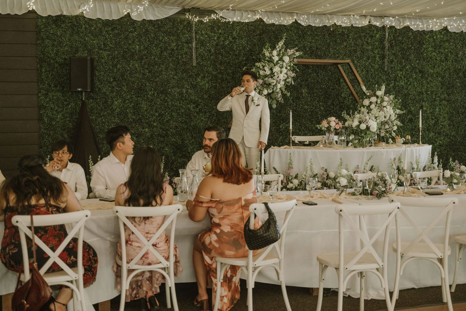 Wedding guests seated at reception tables while groomsman delivers toast near floral head table.