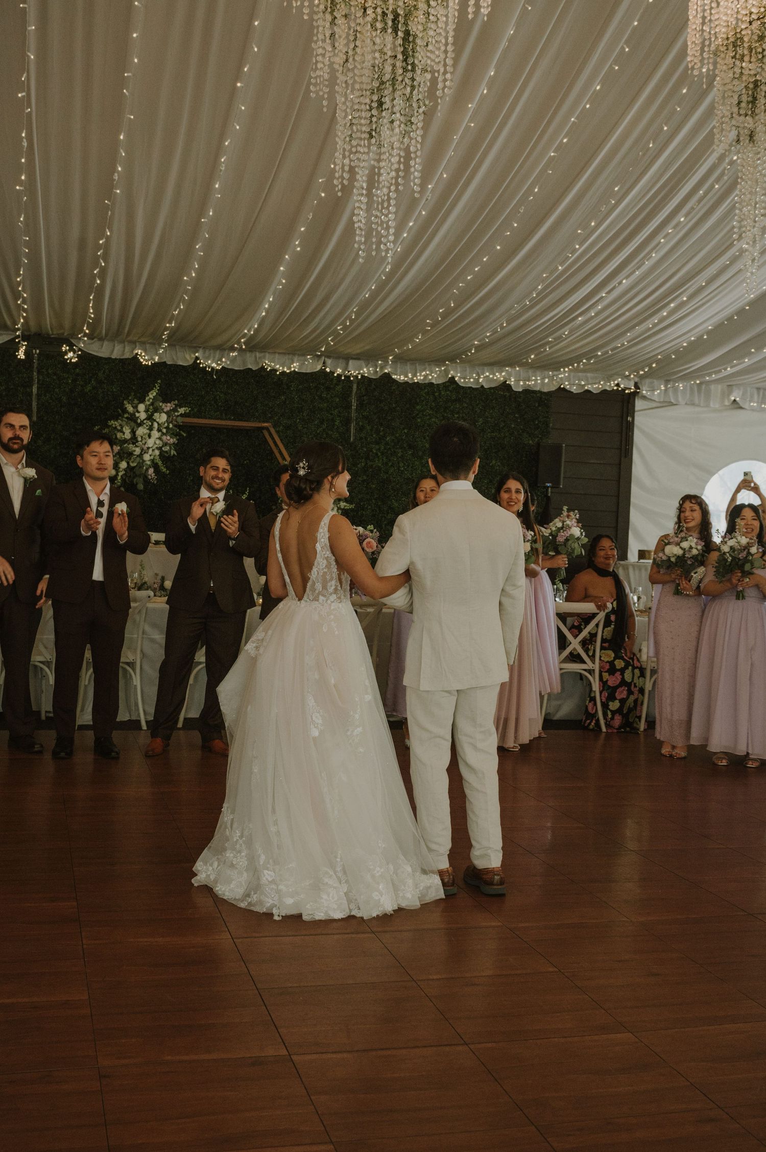 Bride and groom share first dance under draped white ceiling with fairy lights at elegant wedding reception.