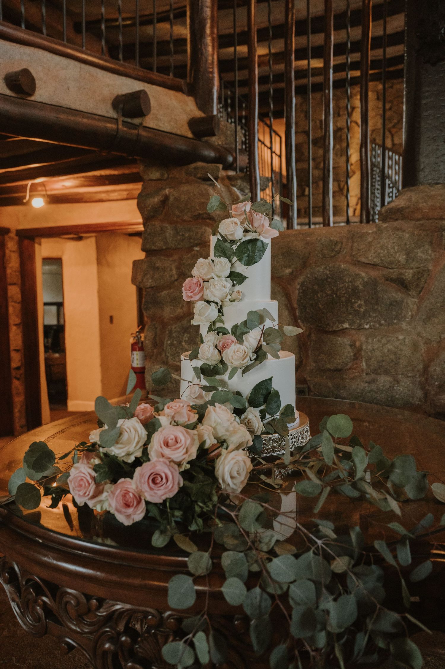 Elegant white wedding cake adorned with blush pink roses and eucalyptus greenery displayed on a wooden table in a rustic stone venue.