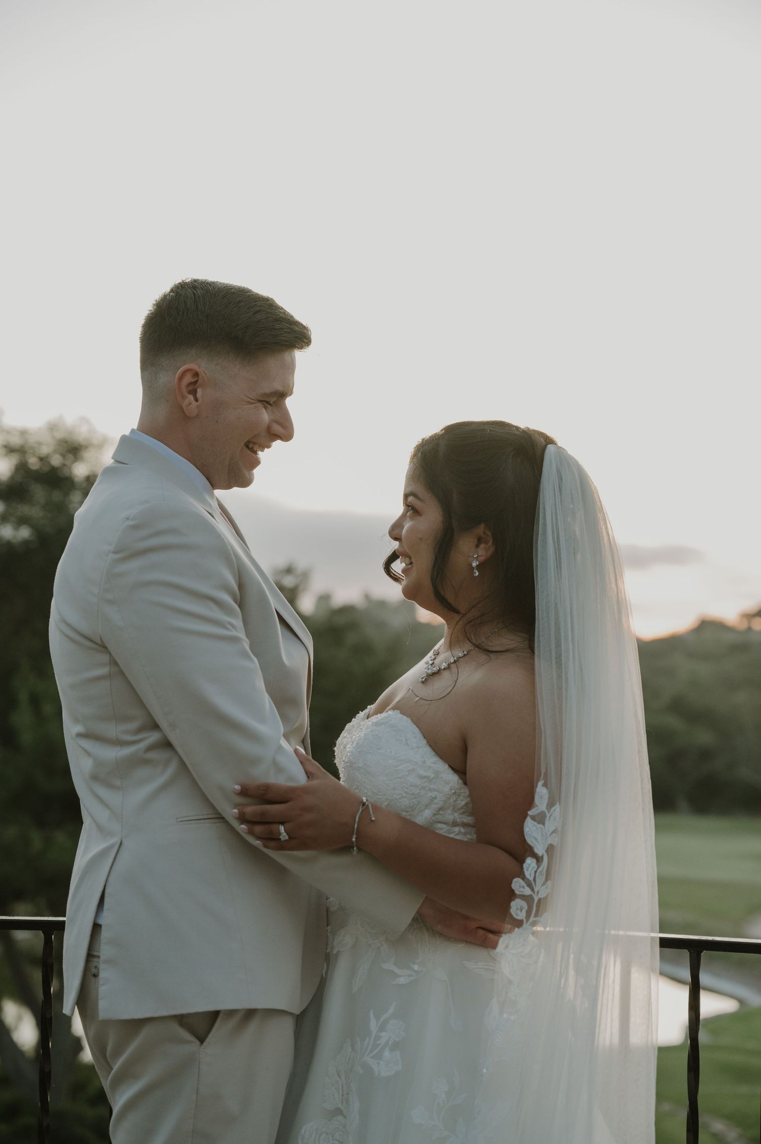 Bride in white lace gown with veil and groom in beige suit sharing romantic sunset moment on balcony.