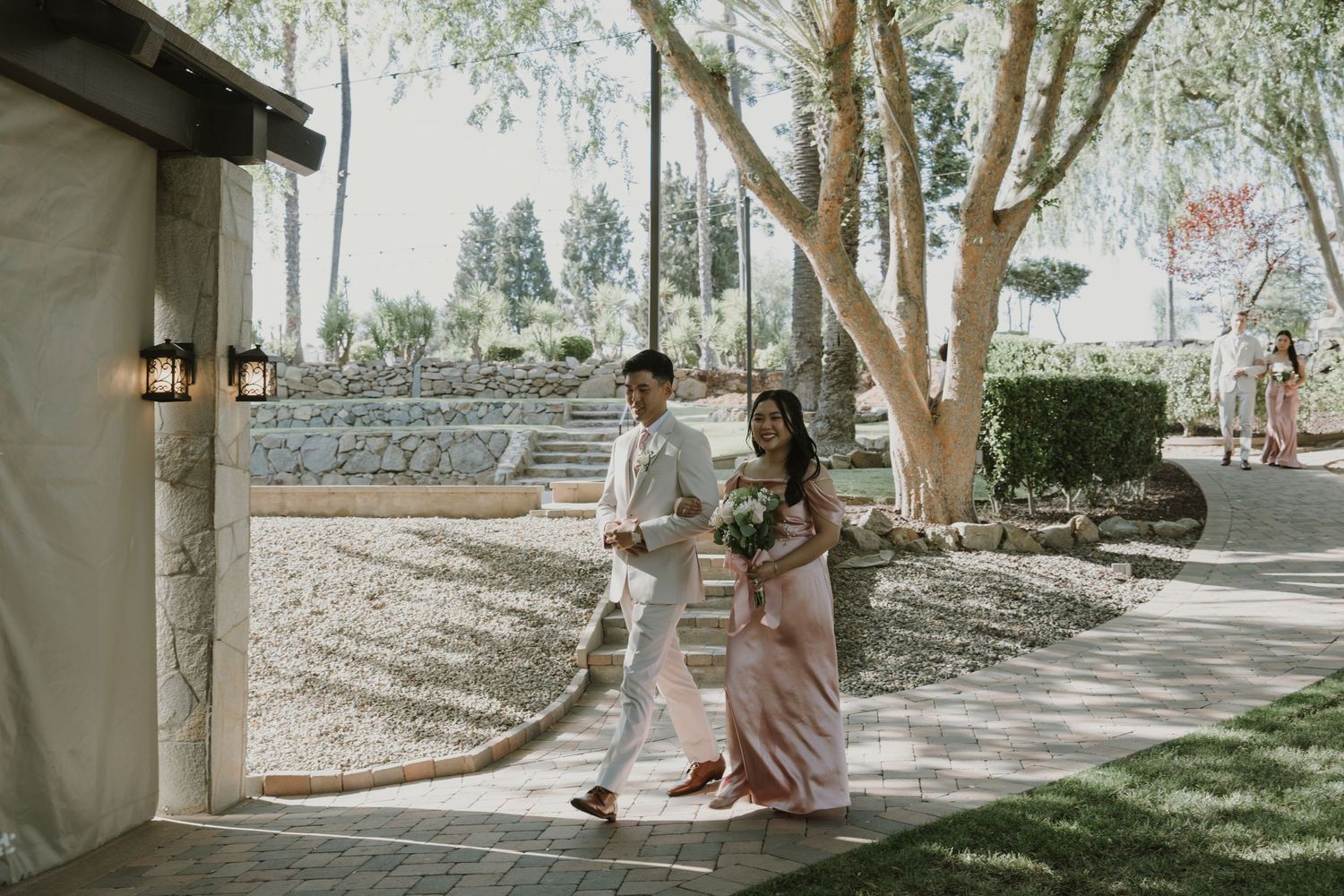 Bride and groom walking together through an elegant outdoor wedding venue with lush trees and stone landscaping.