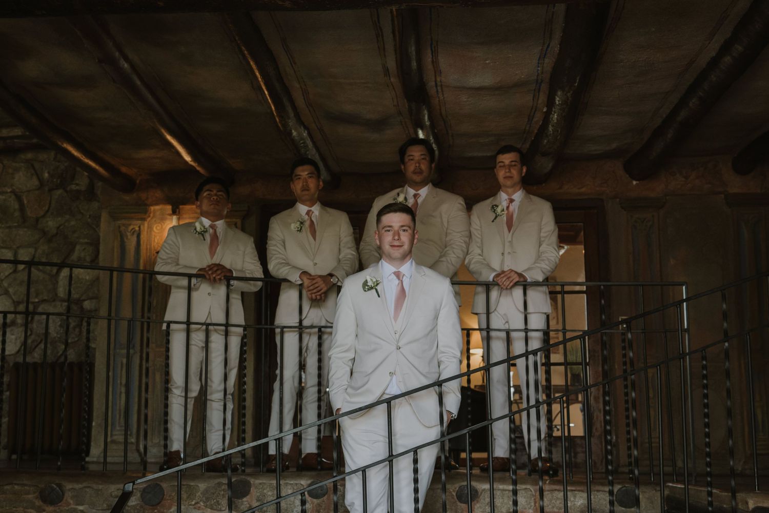 Groom in white suit stands before groomsmen in tan suits on a rustic stone balcony at a wedding venue.