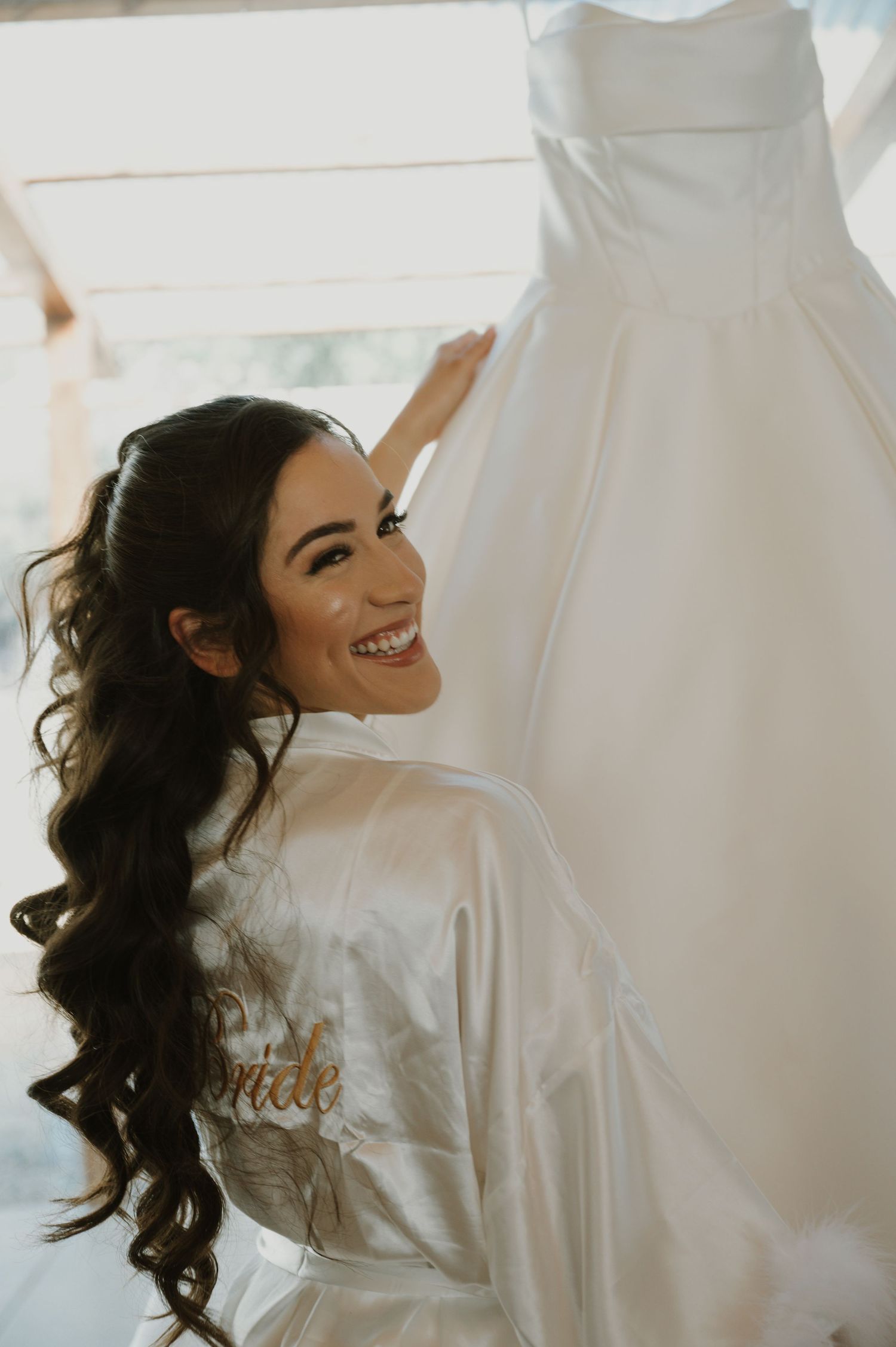 Bride in white silk robe smiles joyfully while getting ready on wedding morning.