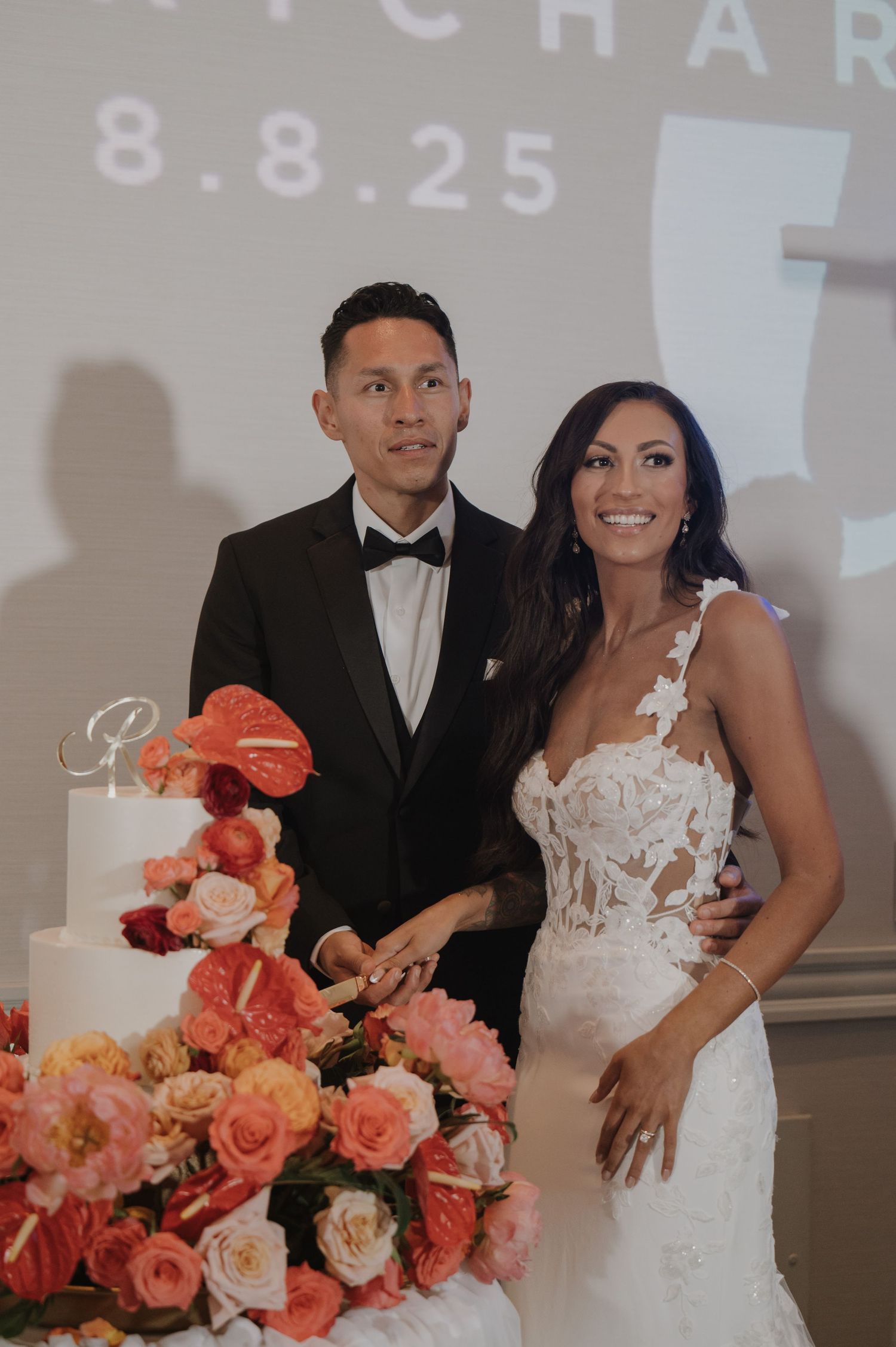 Wedding couple poses by coral rose floral arrangements in elegant venue setting.