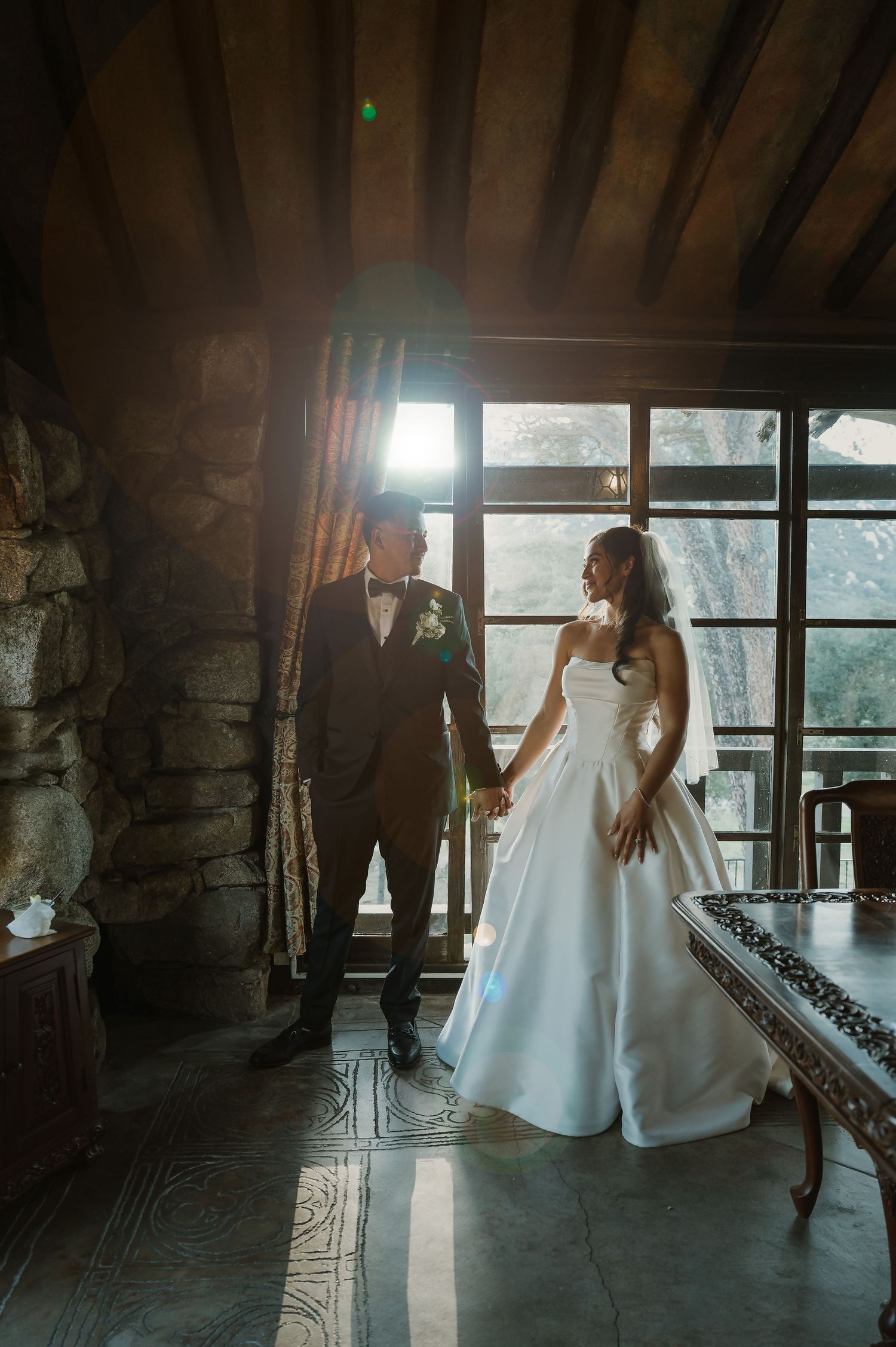 Sunlit wedding portrait in rustic stone room with large windows.