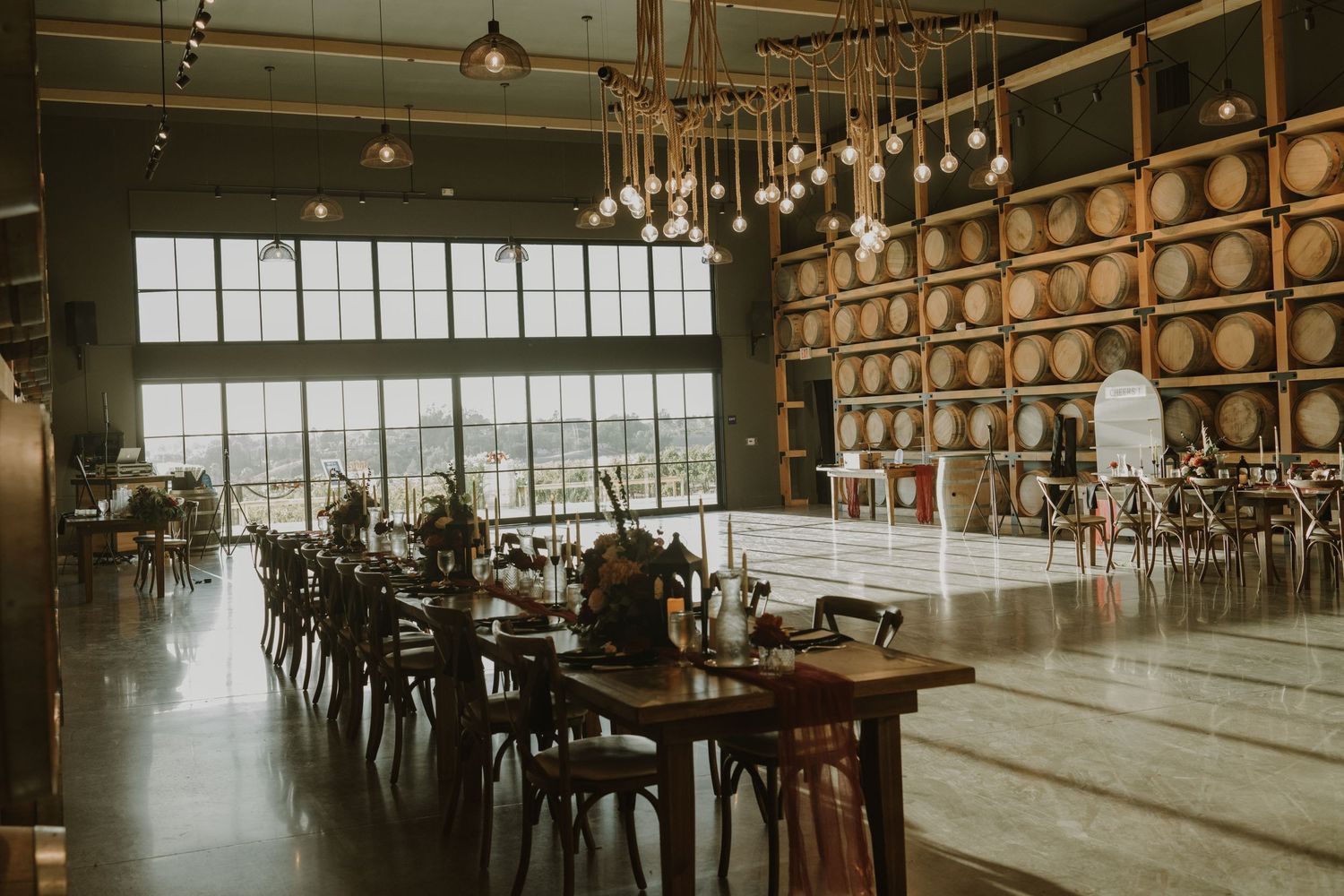 Modern winery event space with floor-to-ceiling windows, barrel walls, and pendant lighting.