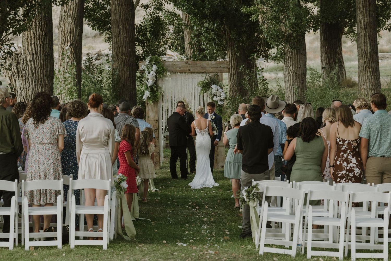 White folding chairs line a shaded outdoor wedding venue with guests arriving for the ceremony.