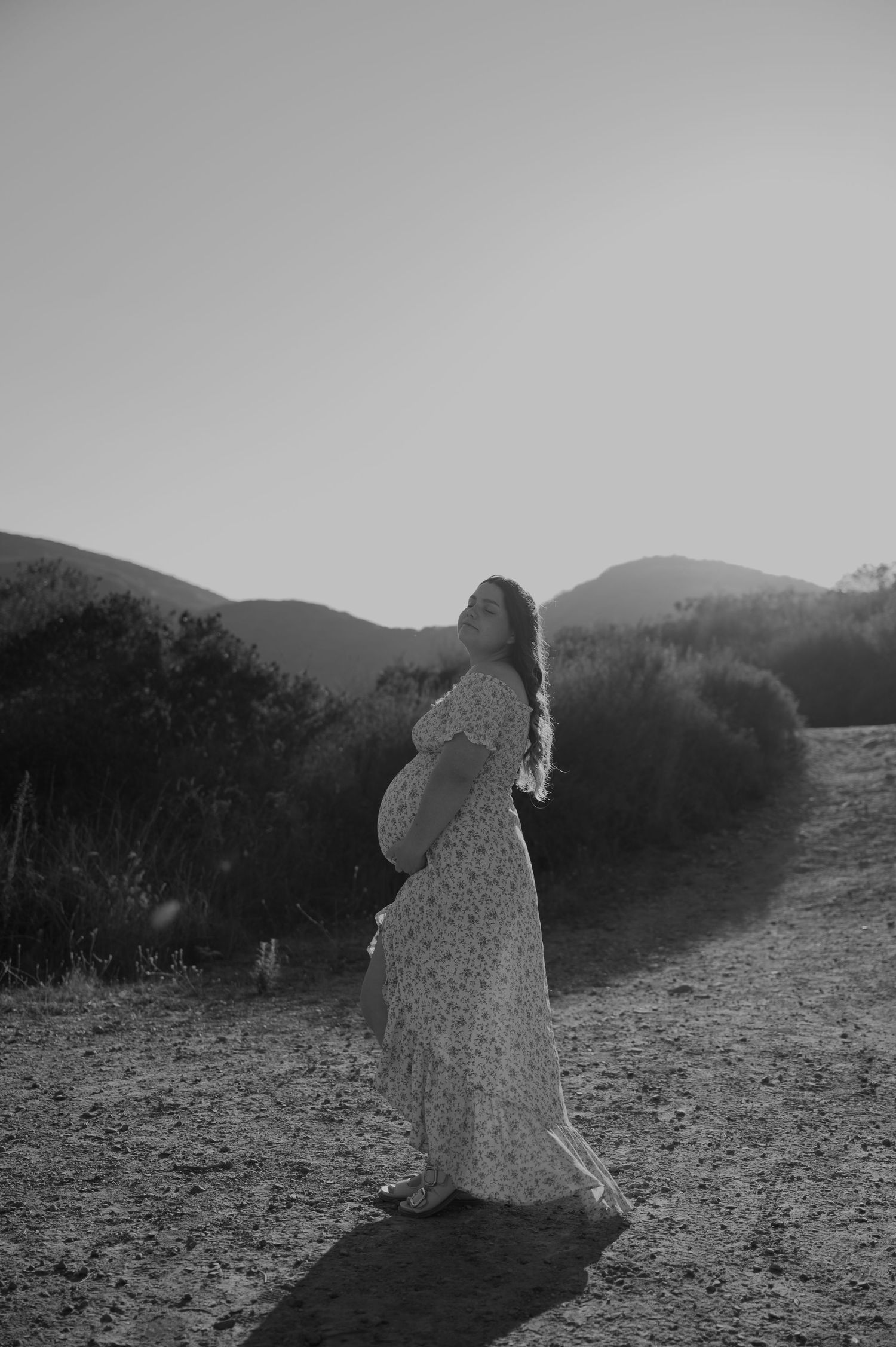 A silhouette of an expectant mother stands against rolling hills in black and white.