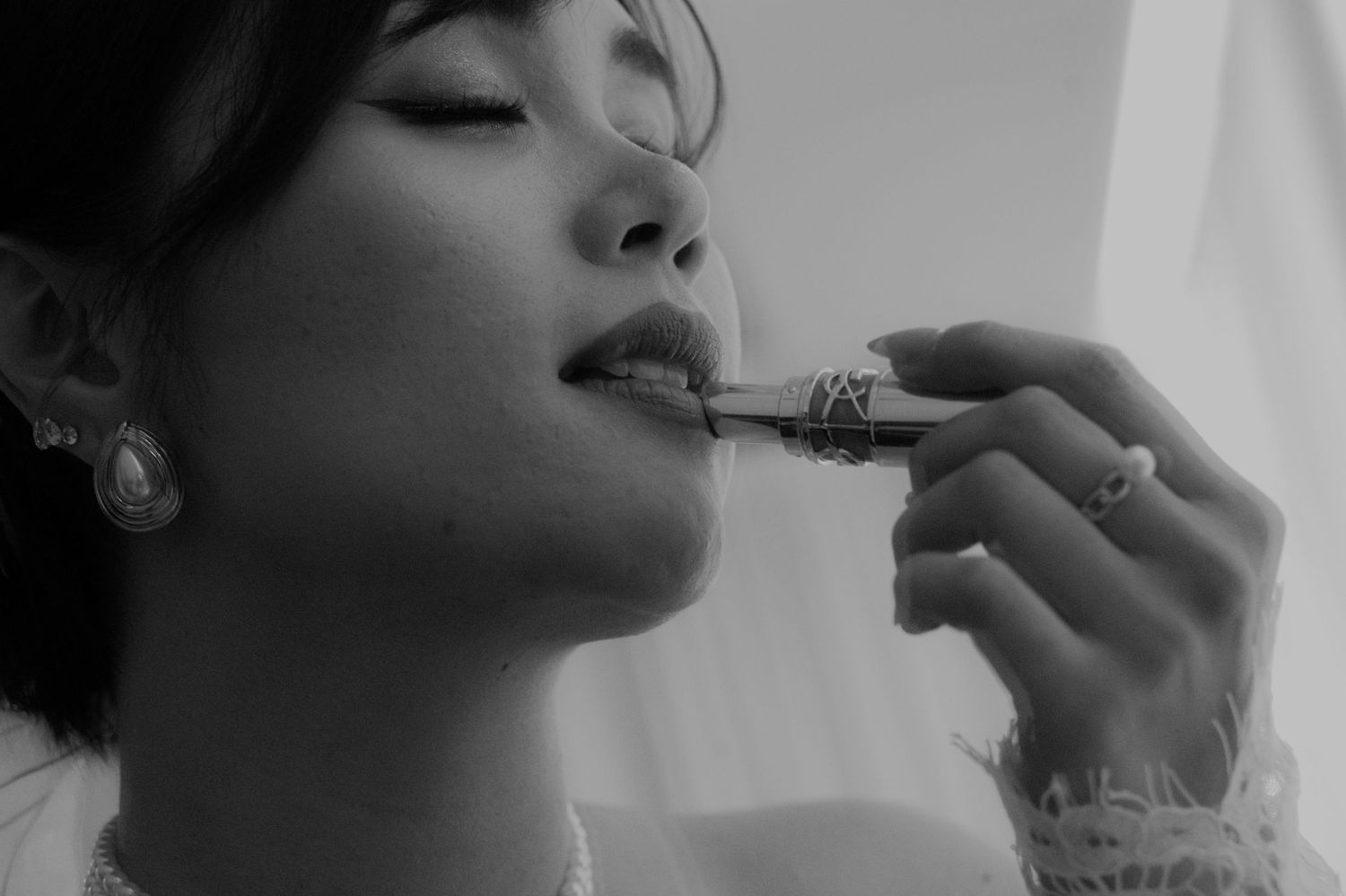 Artistic black and white close-up of woman applying lipstick while wearing elegant jewelry.
