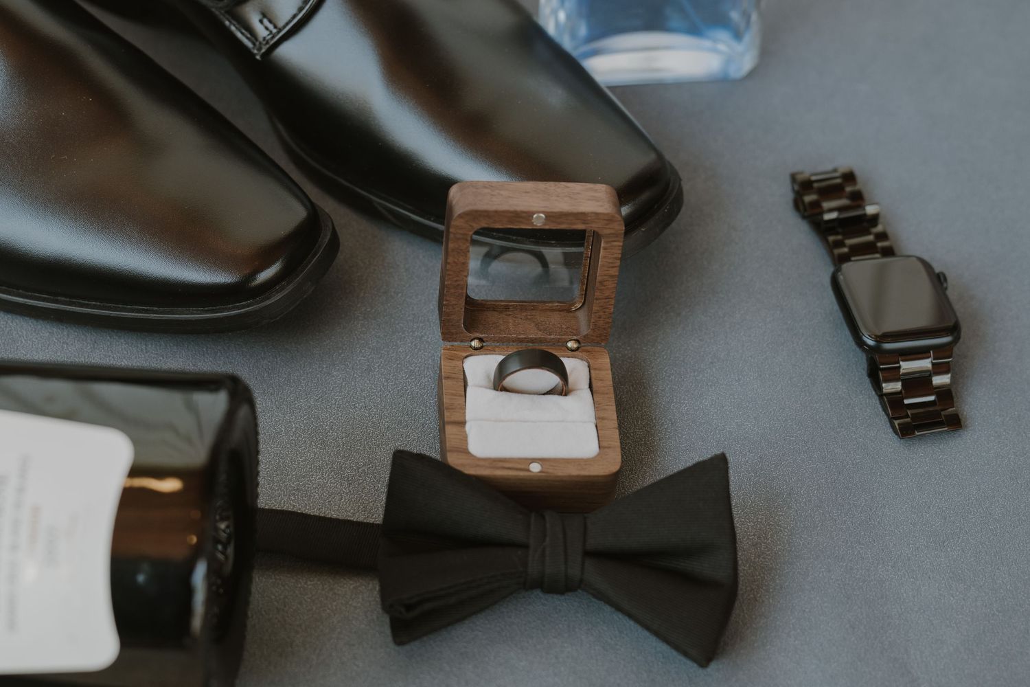 Groom wedding flat lay with black dress shoes, bow tie, ring box, watch, and cologne on gray surface.