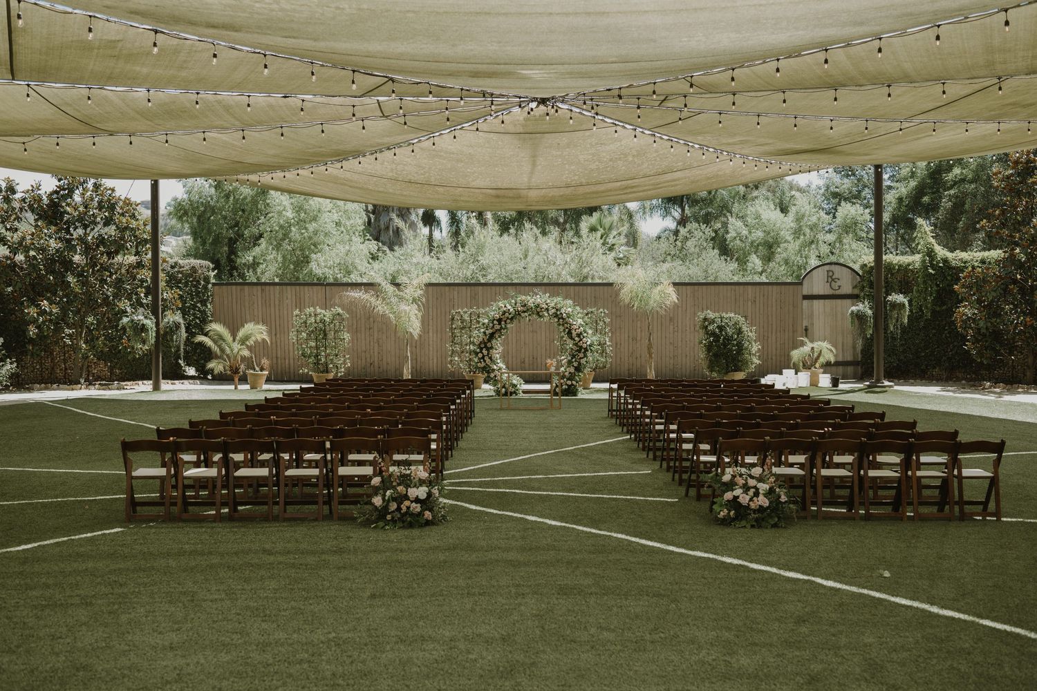 Outdoor wedding ceremony setup with rows of wooden chairs on green grass under a draped canopy with string lights and floral arch.