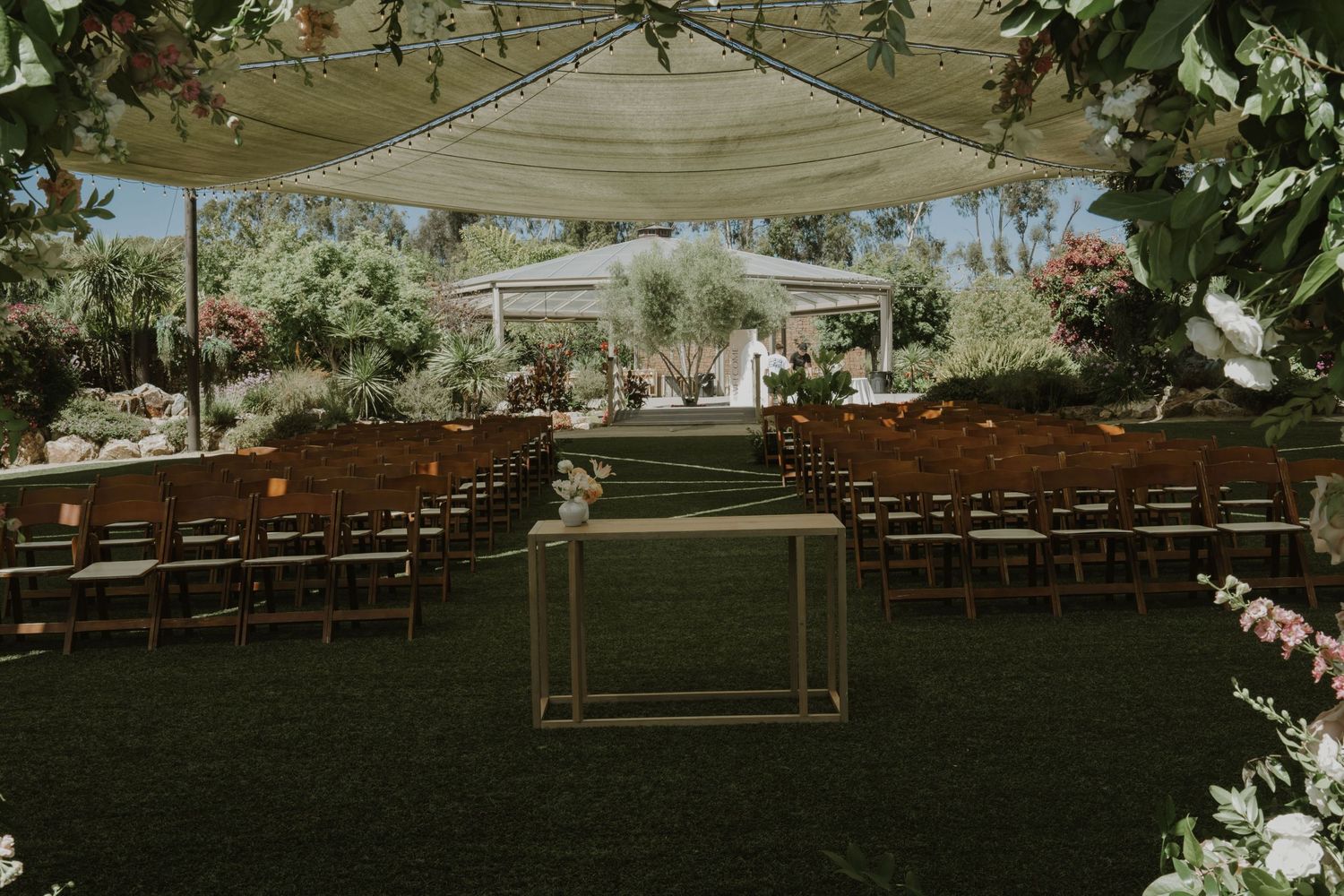 Elegant outdoor wedding ceremony setup with wooden chairs, floral arrangements, and a white canopy on lush green lawn.