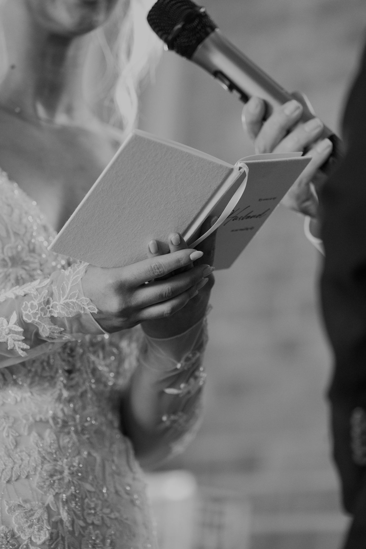 Bride reading wedding vows from a booklet while holding a microphone, wearing a lace gown, black and white photo.
