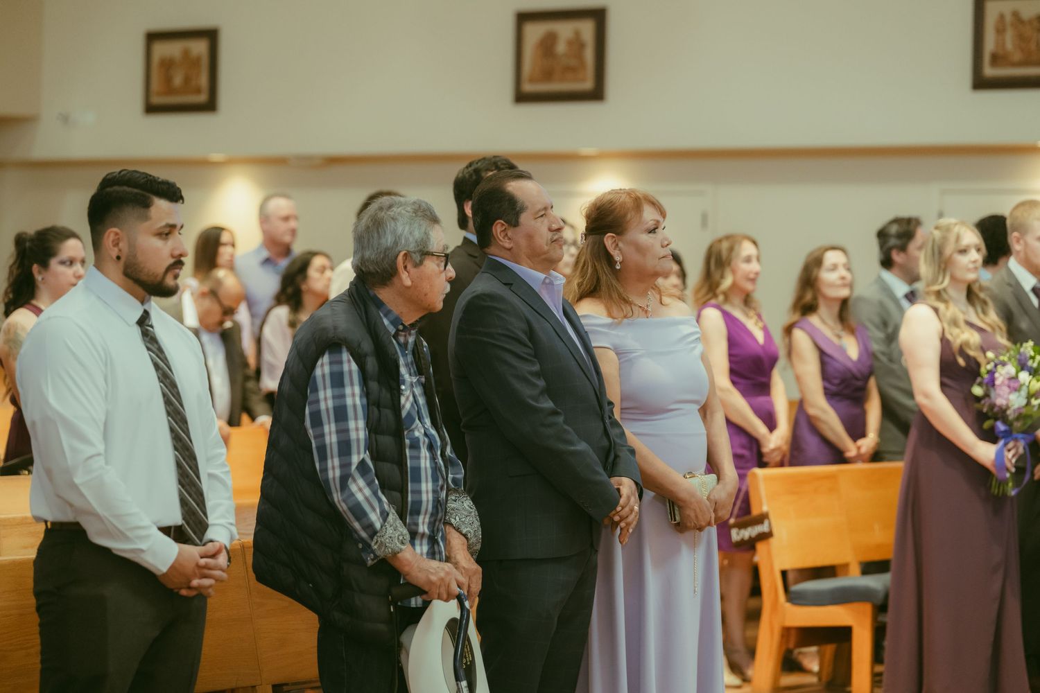 Guests standing in church pews during a wedding ceremony, with bridesmaids in purple dresses visible on the right.