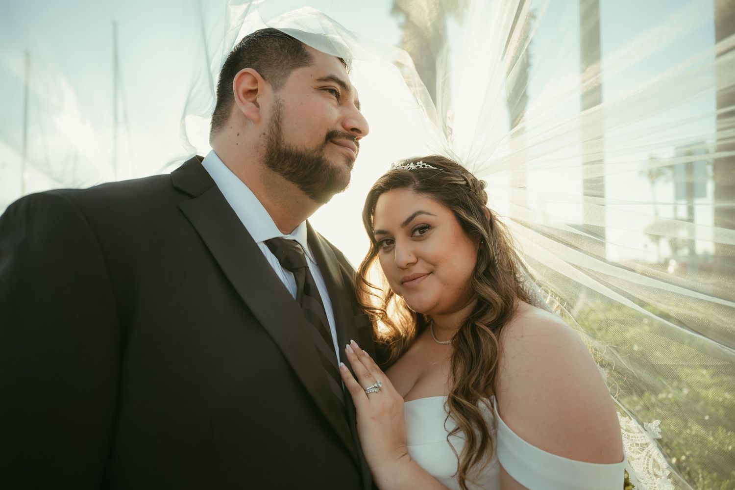 Bride and groom posing outdoors in golden sunlight, groom in black suit, bride in white dress with floral hair accessory.