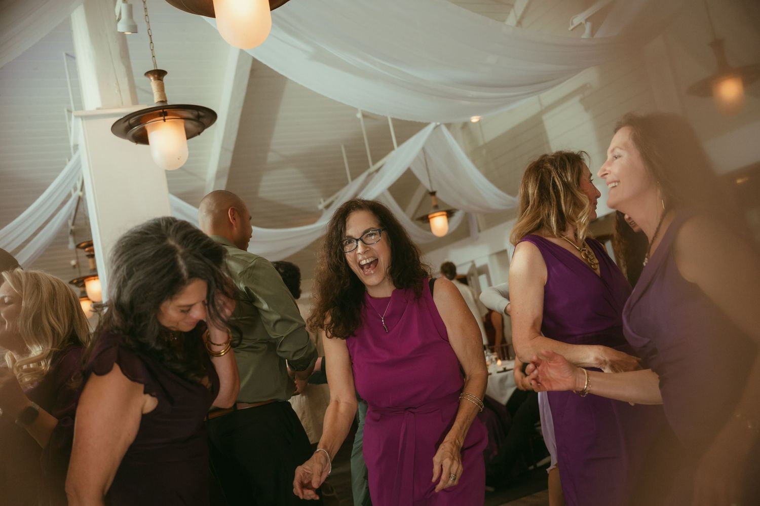 Women in purple dresses laughing and dancing together at an elegant wedding reception with draped ceiling decor.