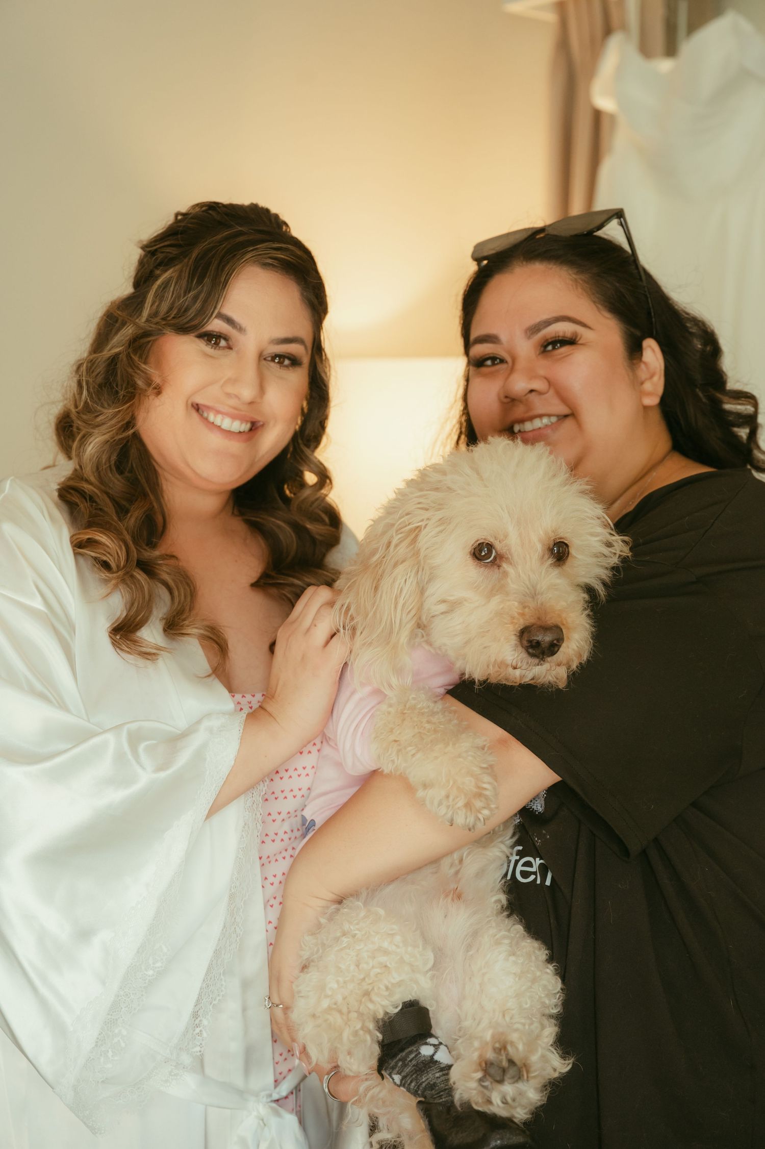 Bride in white robe smiles with friend holding fluffy white dog in pink outfit during wedding preparations.