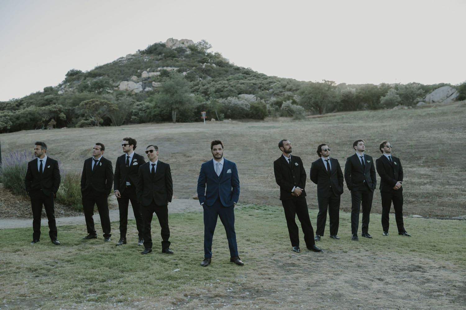 A groom in a blue suit stands centered among groomsmen in black suits on a grassy hillside.
