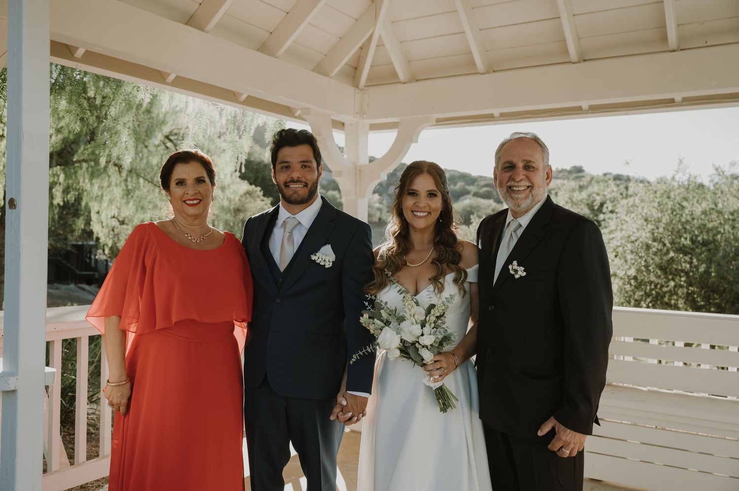 A wedding couple poses with two family members under a white gazebo at an outdoor venue.