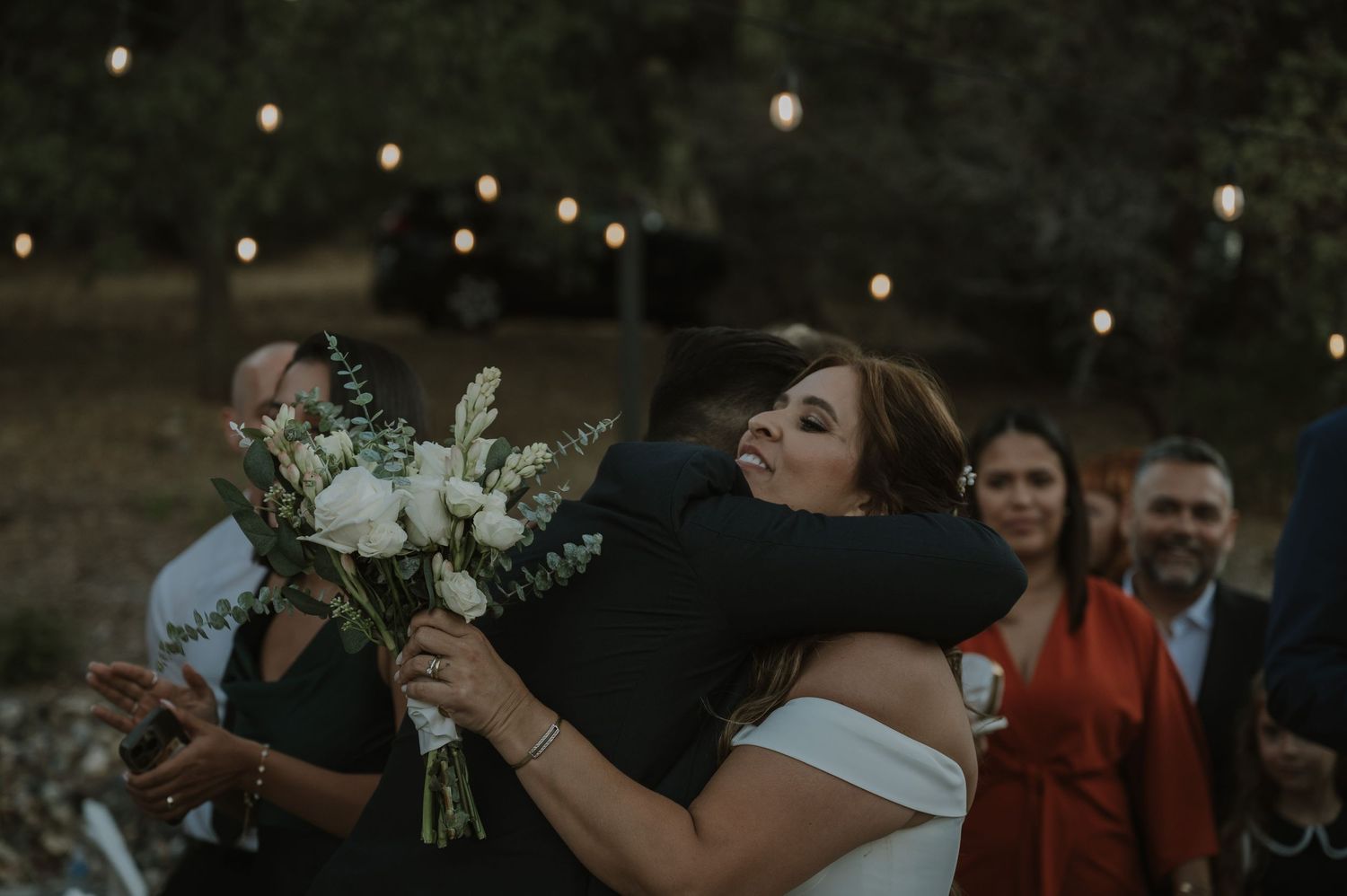 Bride and groom share a heartfelt hug at outdoor wedding ceremony with white bouquet and string lights.