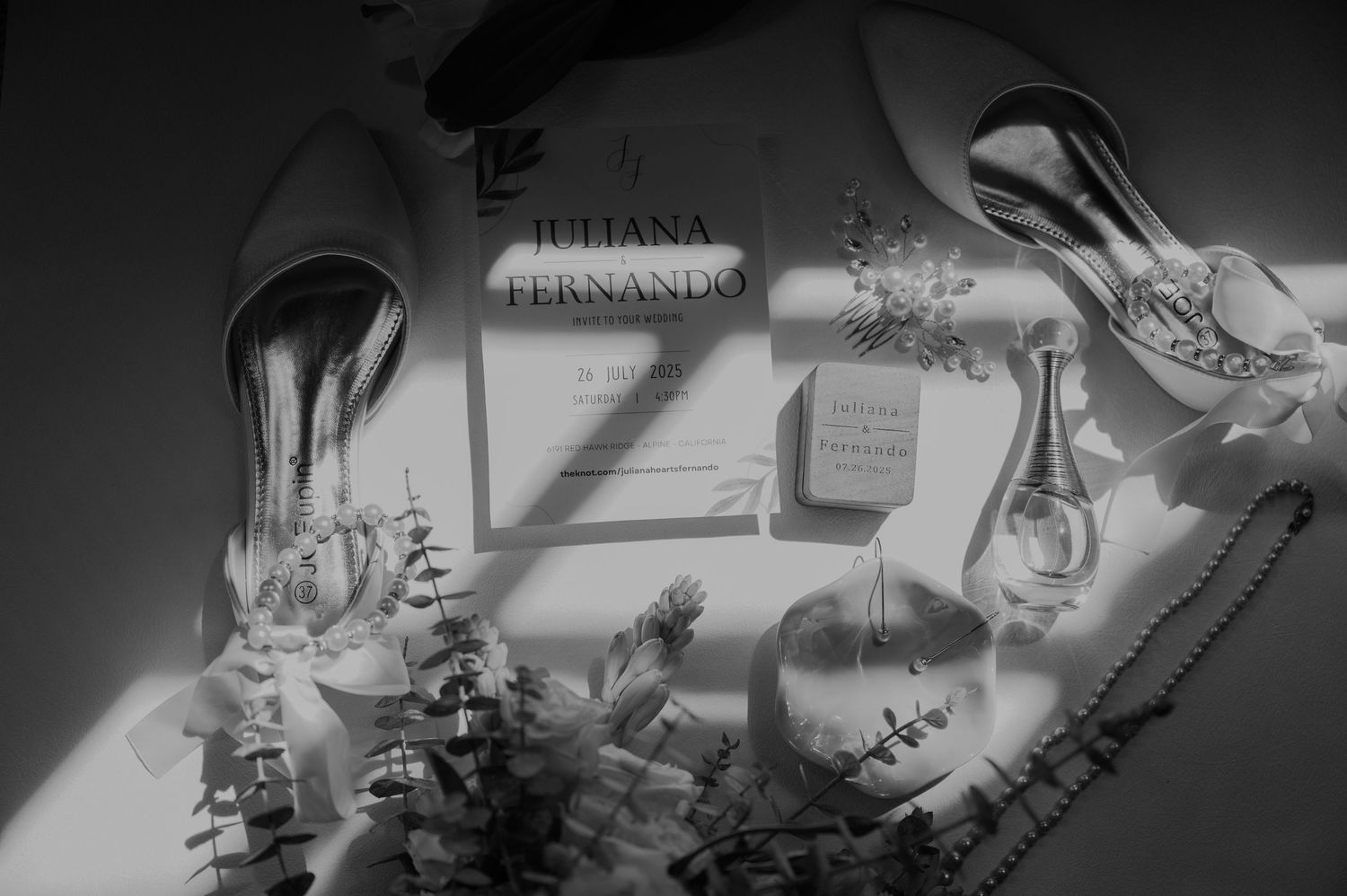 Black and white flat lay of wedding details including heels, invitation, perfume, and pearls.