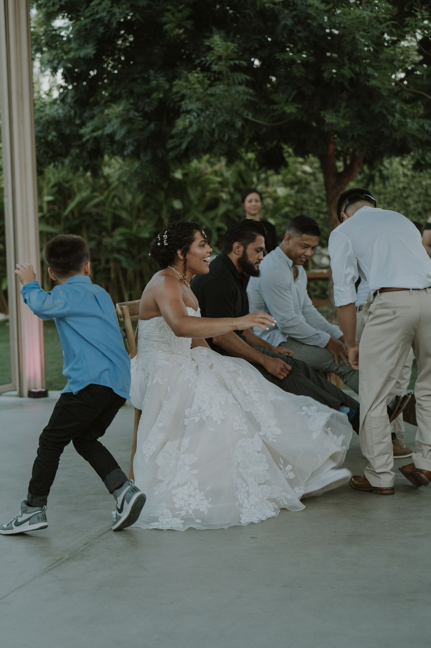 Bride in white gown laughing as guests playfully lift her chair during outdoor wedding reception.
