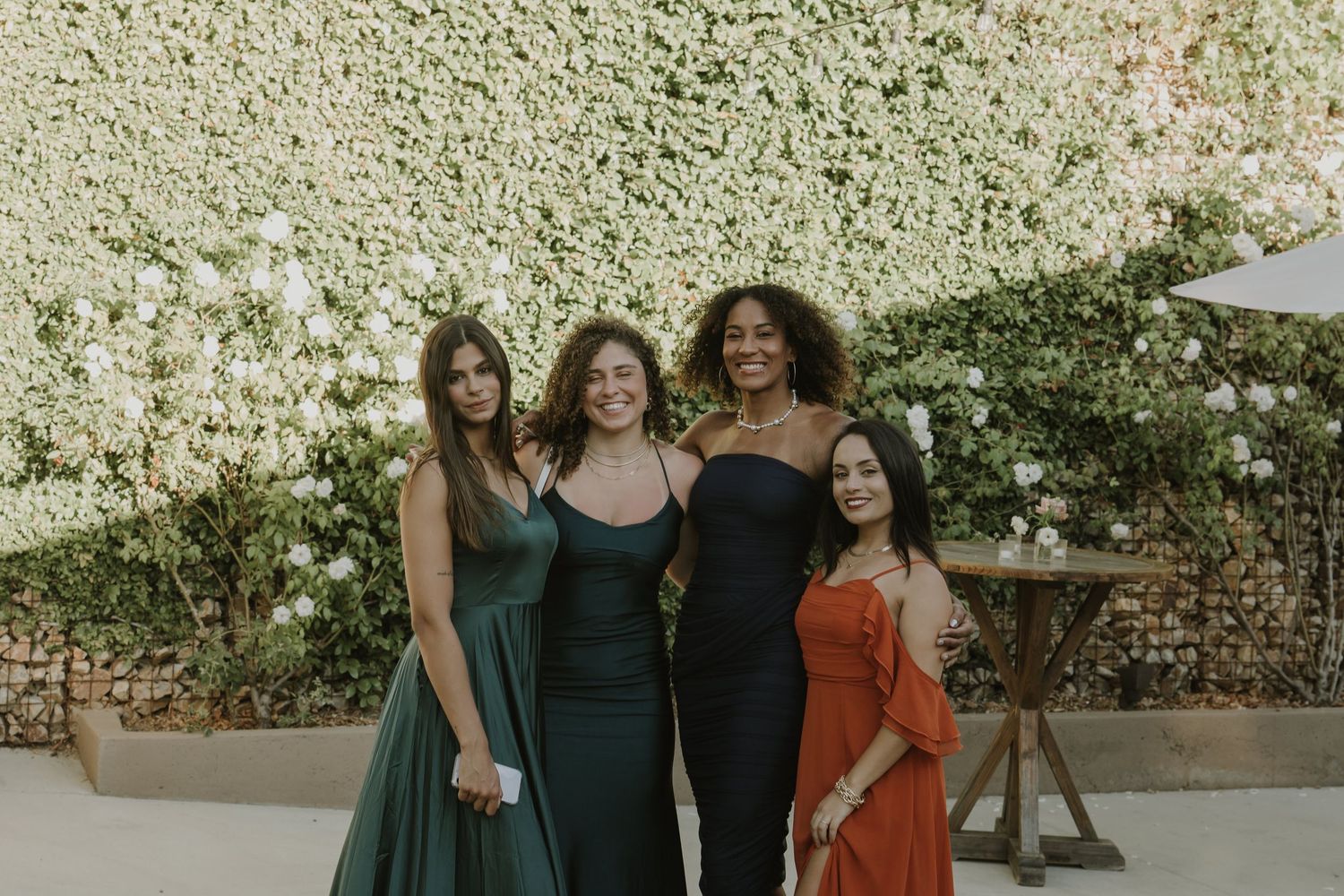 Four women in elegant dresses posing together in front of a lush floral wall backdrop.