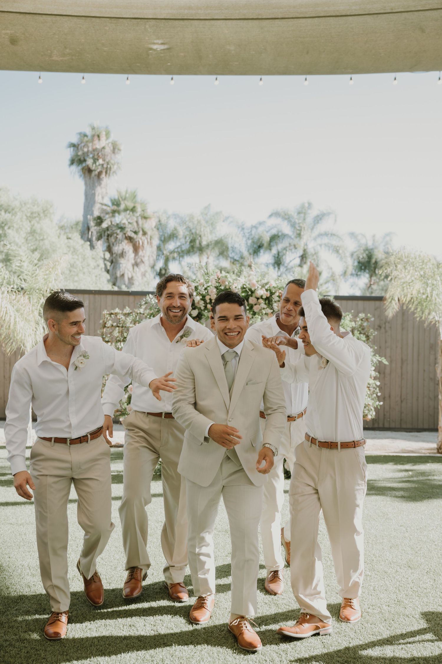 Groom celebrates with groomsmen in tan suits and white shirts, laughing outdoors near a floral arch backdrop.