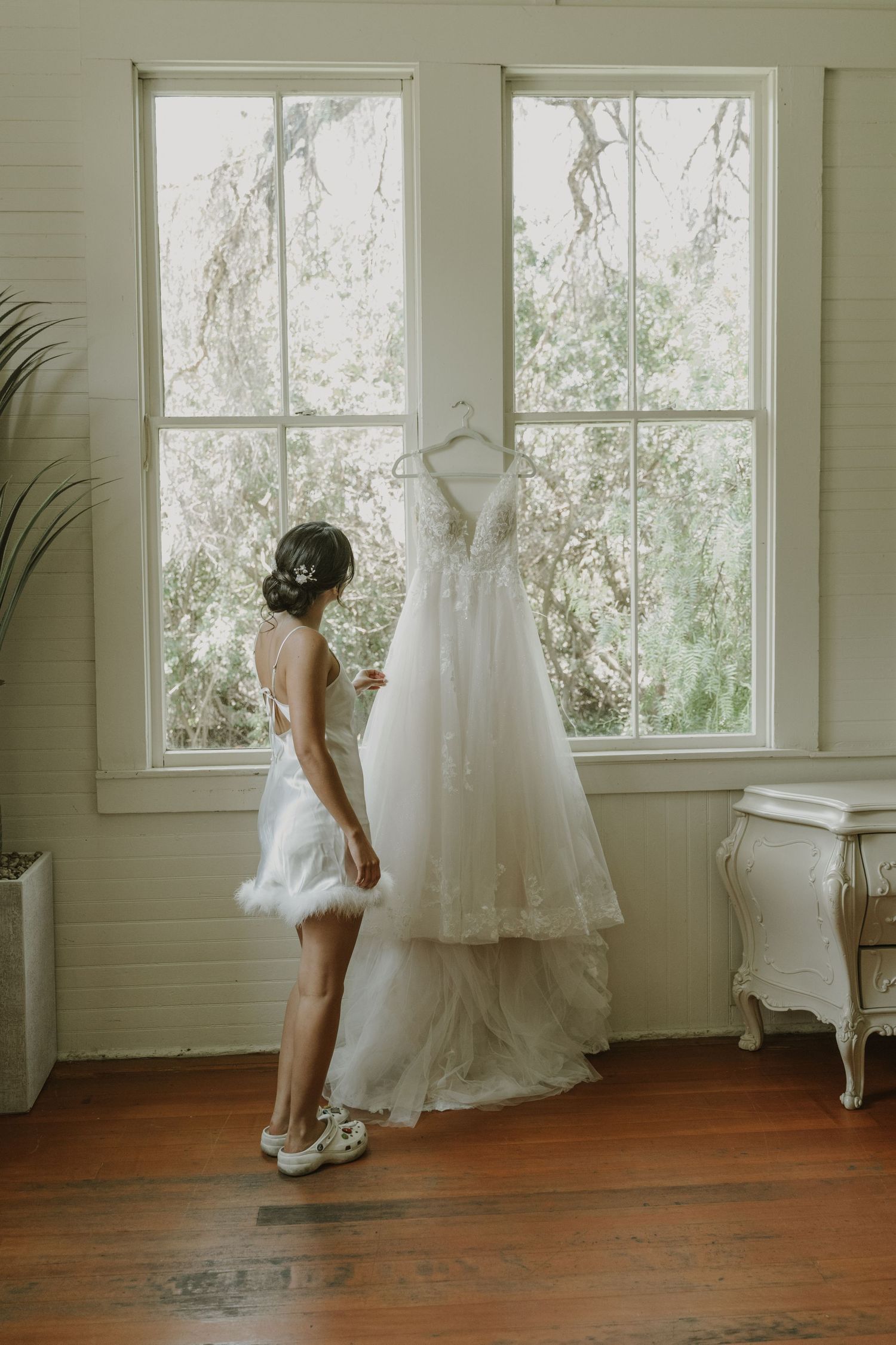 Bride in white robe admiring her lace wedding gown hanging by a sunlit window in a vintage room.