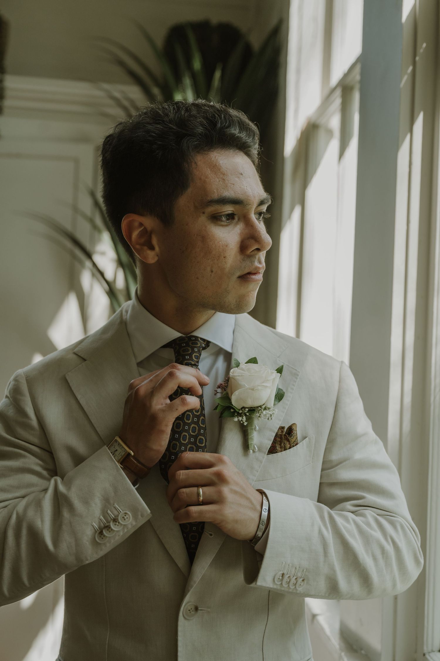 Groom in a beige suit adjusting his patterned tie with a white rose boutonniere near a bright window.