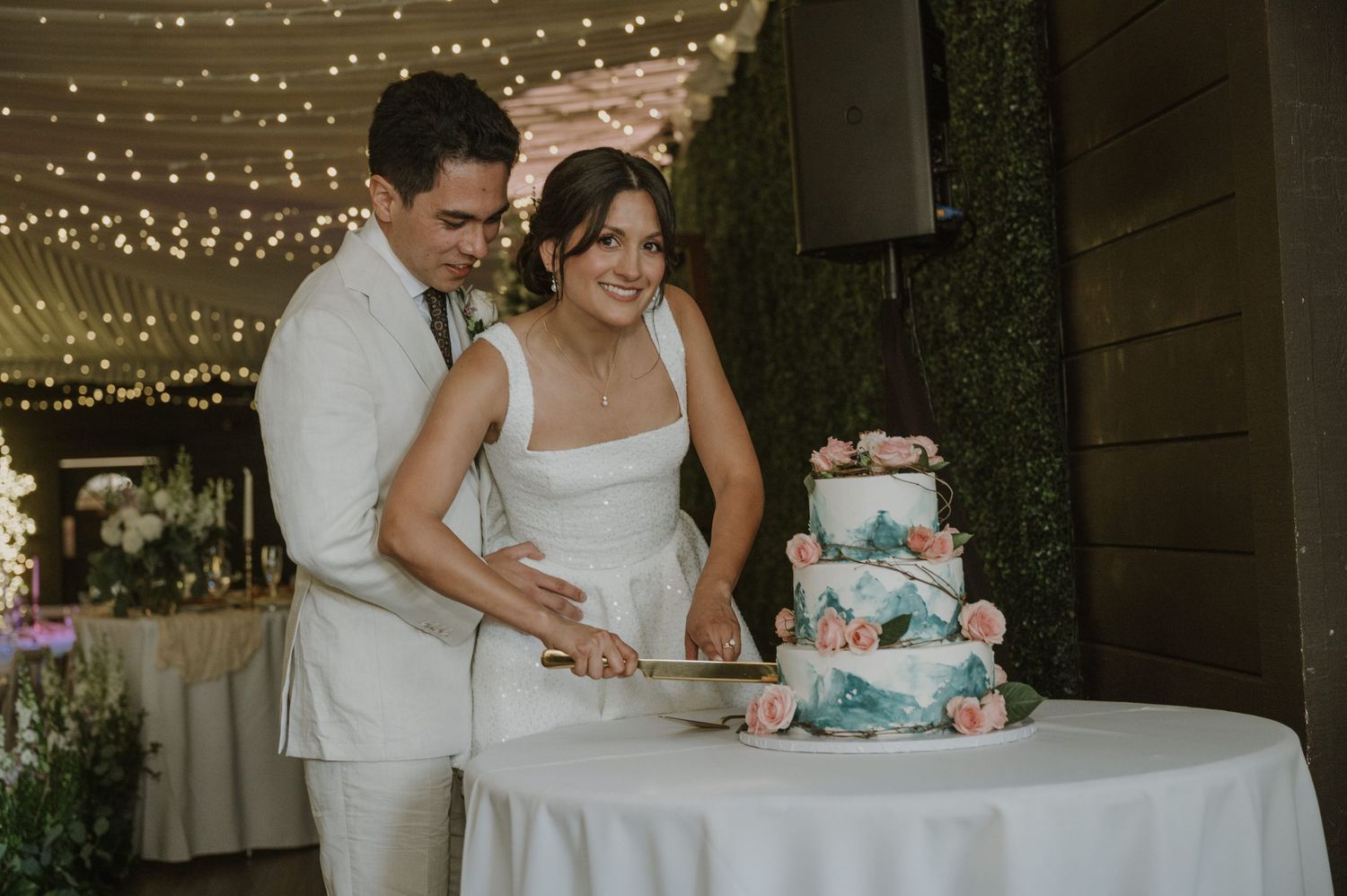 Bride and groom cut elegant three-tier floral wedding cake under warm fairy lights.