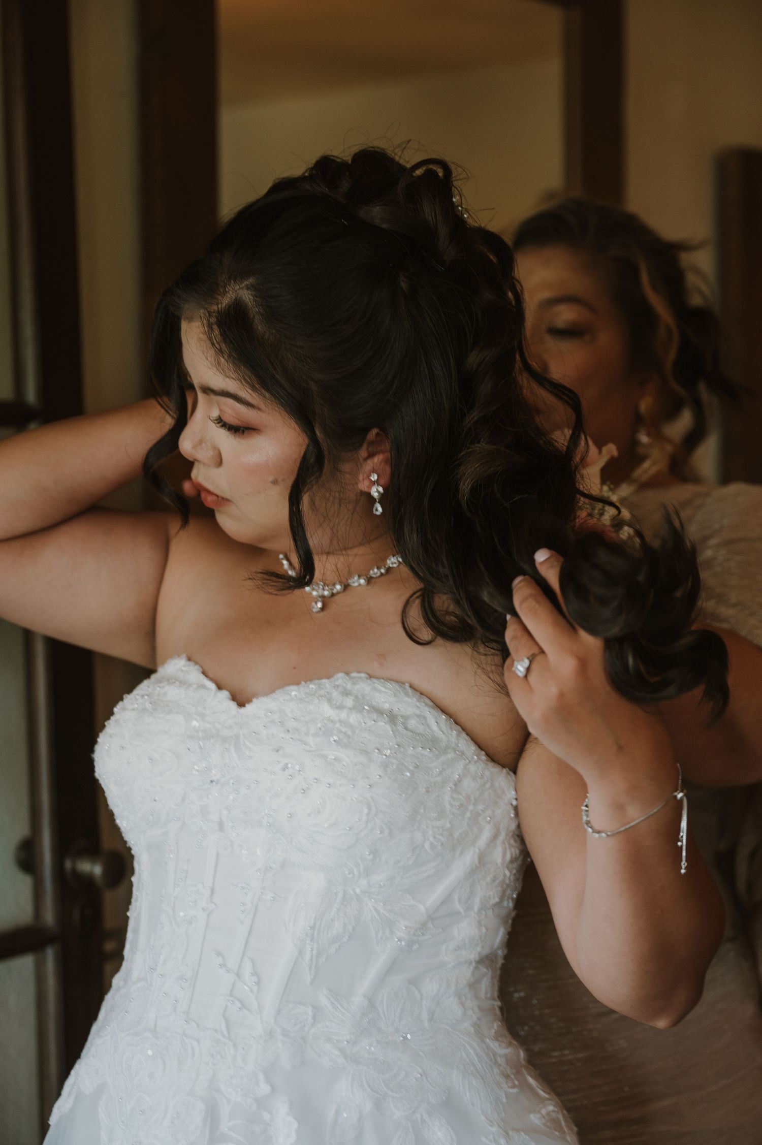 Bride in strapless lace wedding gown getting hair styled by bridesmaid, wearing diamond necklace and earrings.