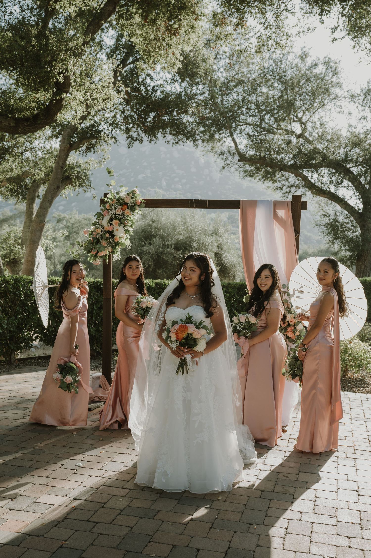 Bride in white gown holds bouquet surrounded by bridesmaids in blush pink dresses under wooden arch outdoors.