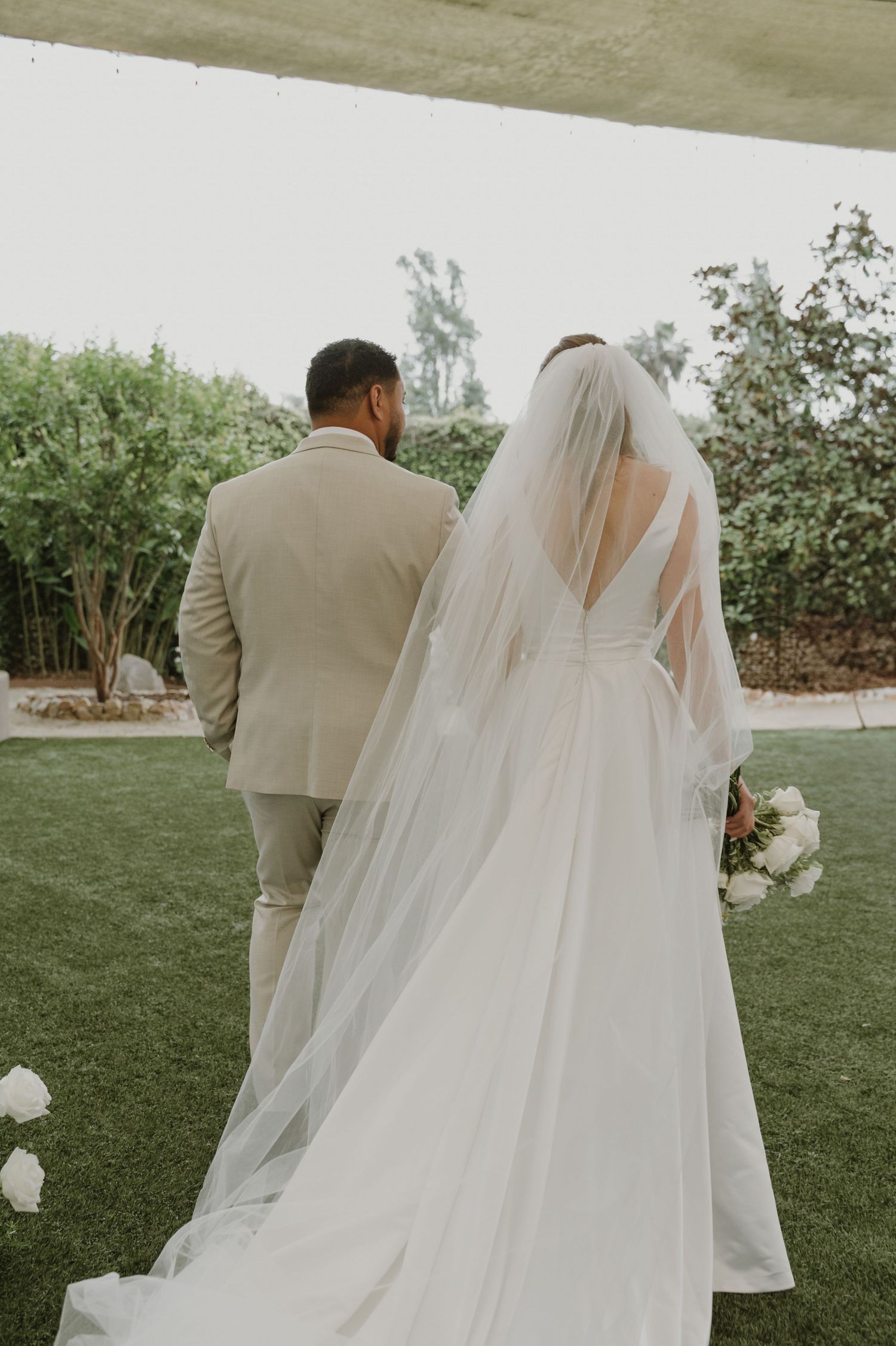 Bride in white gown with veil and groom in tan suit walk hand in hand through lush garden.