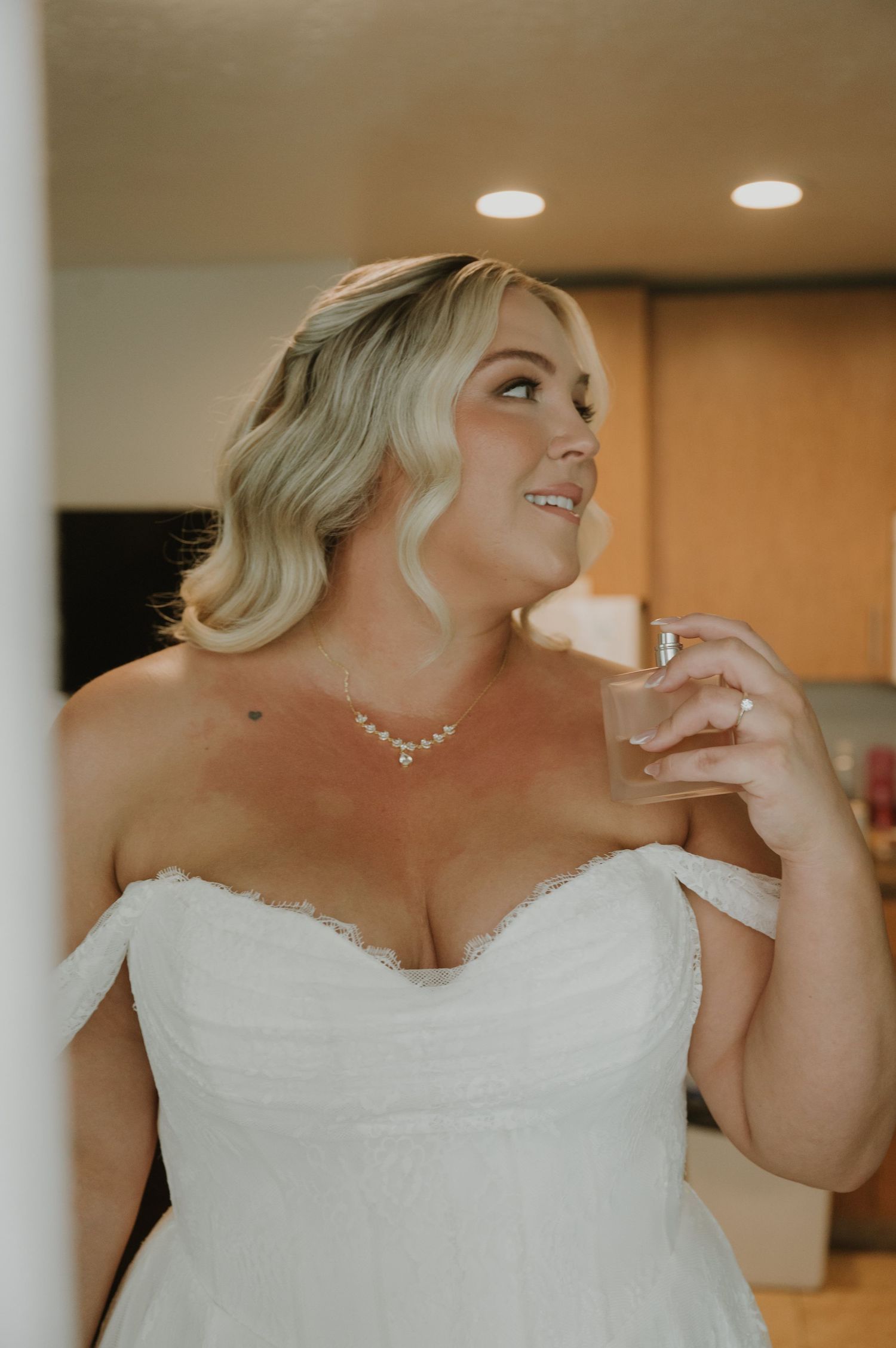 Elegant plus-size bride in off-shoulder white wedding gown applying perfume, wearing pearl necklace with soft blonde waves.
