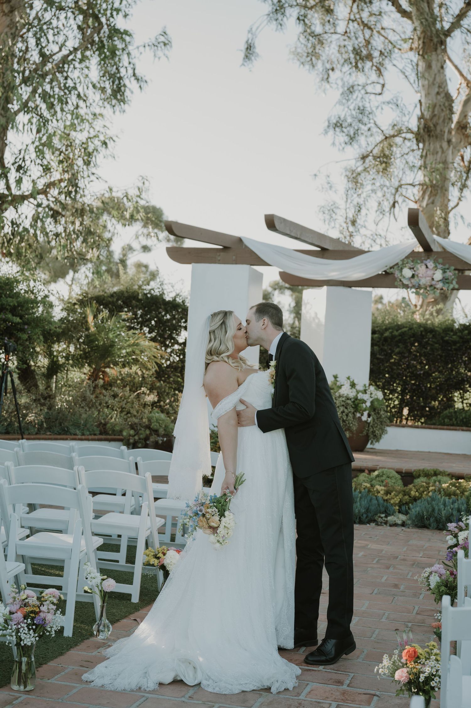 Bride and groom share first kiss at outdoor wedding ceremony beneath wooden arch with white chairs.