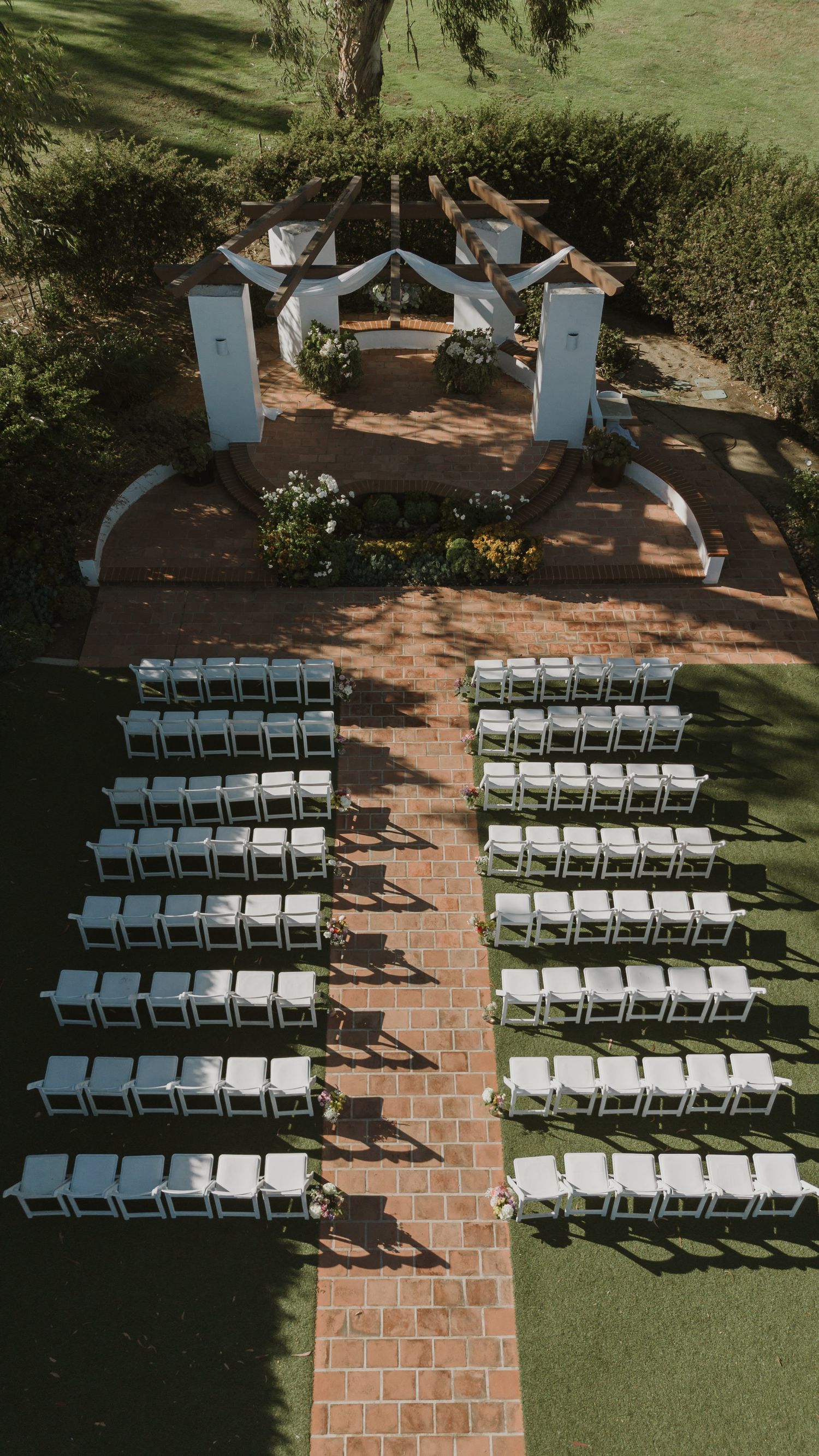 Aerial view of an outdoor wedding ceremony setup with white chairs flanking a brick aisle leading to a white pergola altar.