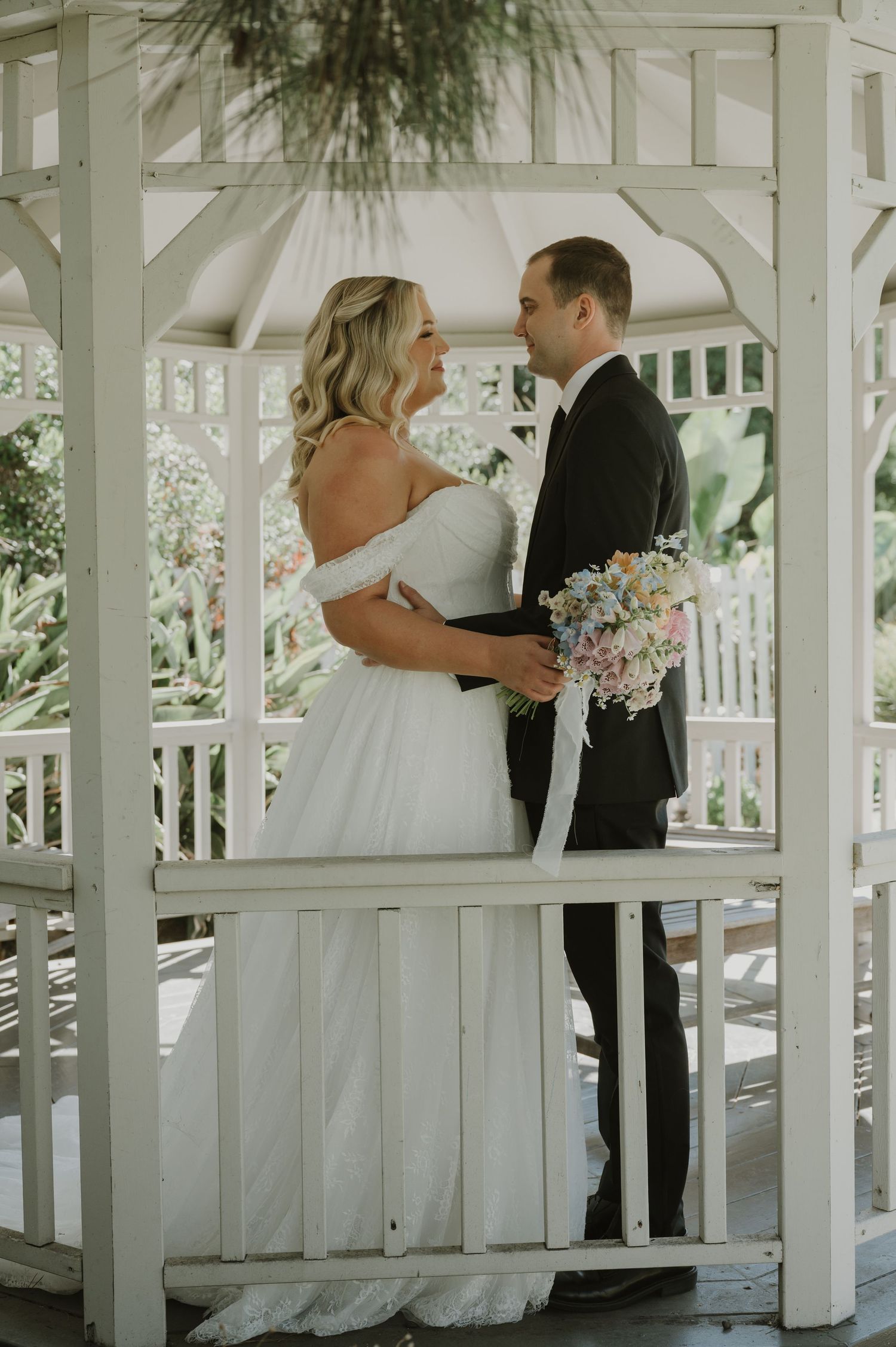 Bride and groom share a romantic moment holding a pastel bouquet inside a white garden gazebo.