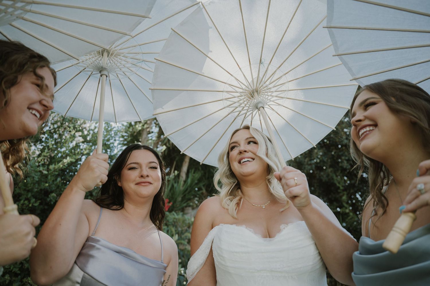 Bride and bridesmaids laugh together under white parasols at an outdoor wedding ceremony.